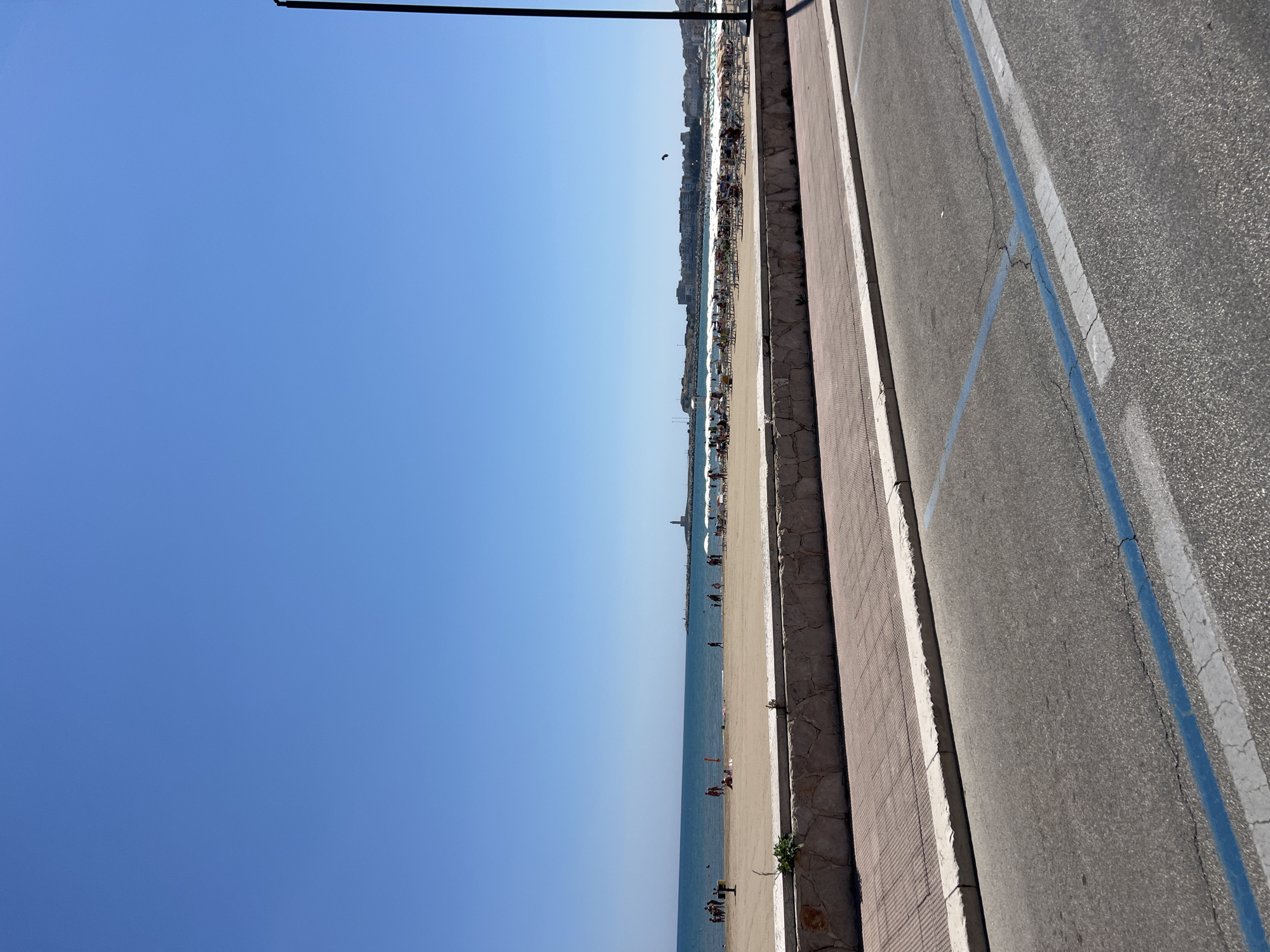 This image depicts a clear, sunny day at a beach in Vieste, Italy. In the foreground, a paved road with blue and white parking lines runs parallel to a pedestrian walkway lined with a stone retaining wall. Beyond the walkway is a sandy beach populated with numerous beachgoers. Many individuals are sunbathing on lounge chairs, some are under white umbrellas, and a few are visible walking or playing near the water's edge. The sea is calm and extends to the horizon, where a distant town skyline with buildings and a lighthouse can be seen. A tall black lamppost stands to the right of the frame, and a small kite is flying in the sky above the town.