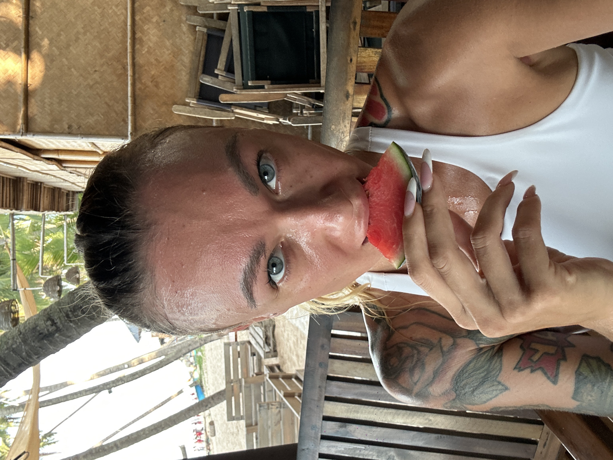 A woman is taking a selfie while eating a slice of watermelon. She is sitting at a table at an outdoor restaurant in Mũi Né, Vietnam. The restaurant appears to be on a beach, with palm trees and the ocean visible in the background. She has tattoos on her left arm and shoulder. The woman is wearing a white tank top.
