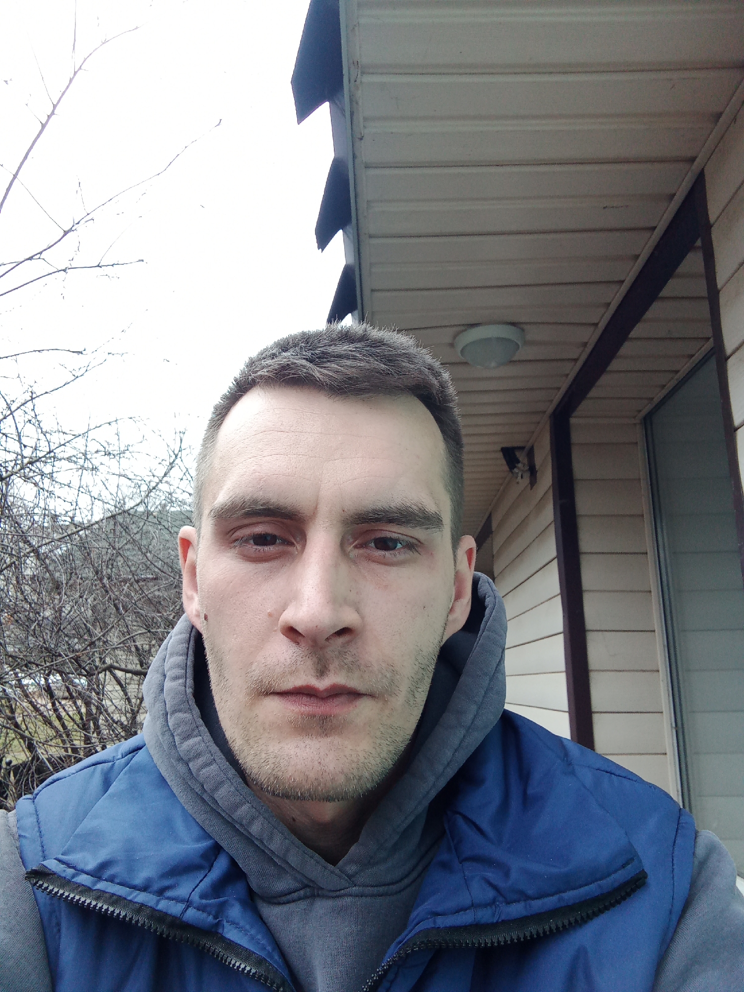 The image shows a head-and-shoulders self-portrait of a fair-skinned man with short brown hair, stubble, and a hoodie under a blue vest. He is outdoors in what appears to be an overcast or cloudy day. The background includes a house with light-colored siding, a bare tree, and possibly a hillside. The location is likely Chaikovsky, Russia.