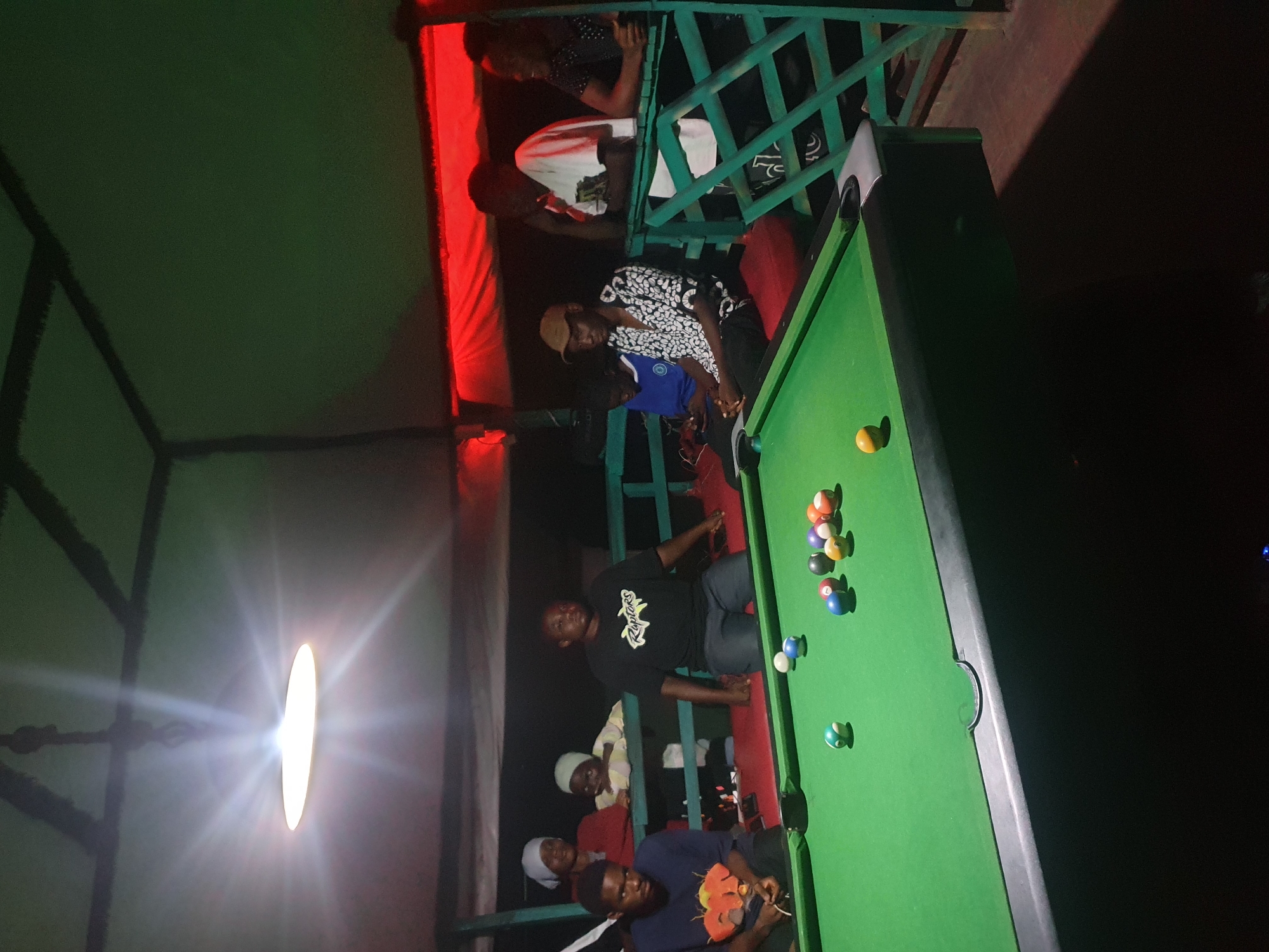 The image depicts a group of young men and women gathered in an outdoor, covered area, likely a bar or lounge, with a pool table as the central focus. The scene is dimly lit, suggesting it is nighttime. Red and green lighting illuminates the space, casting a vibrant glow. A bright, circular light source on the left side of the image creates a lens flare effect.
Several individuals are visible, some actively engaged in playing pool, while others observe or socialize. One man in a black t-shirt with white lettering that appears to say "Raptors" is positioned near the pool table. Another man wears a black and white patterned shirt and a tan baseball cap. A woman in a white top is visible in the background, and others are seen in various states of engagement.
The overall atmosphere appears casual and social. The presence of a pool table and the gathering of people suggest a recreational activity or social event. The setting seems to be an elevated or multi-level structure, indicated by the railings and the way people are positioned on different levels. The red lighting, railing structure, and clothing styles hint at a location in Nigeria, specifically Lokoja.