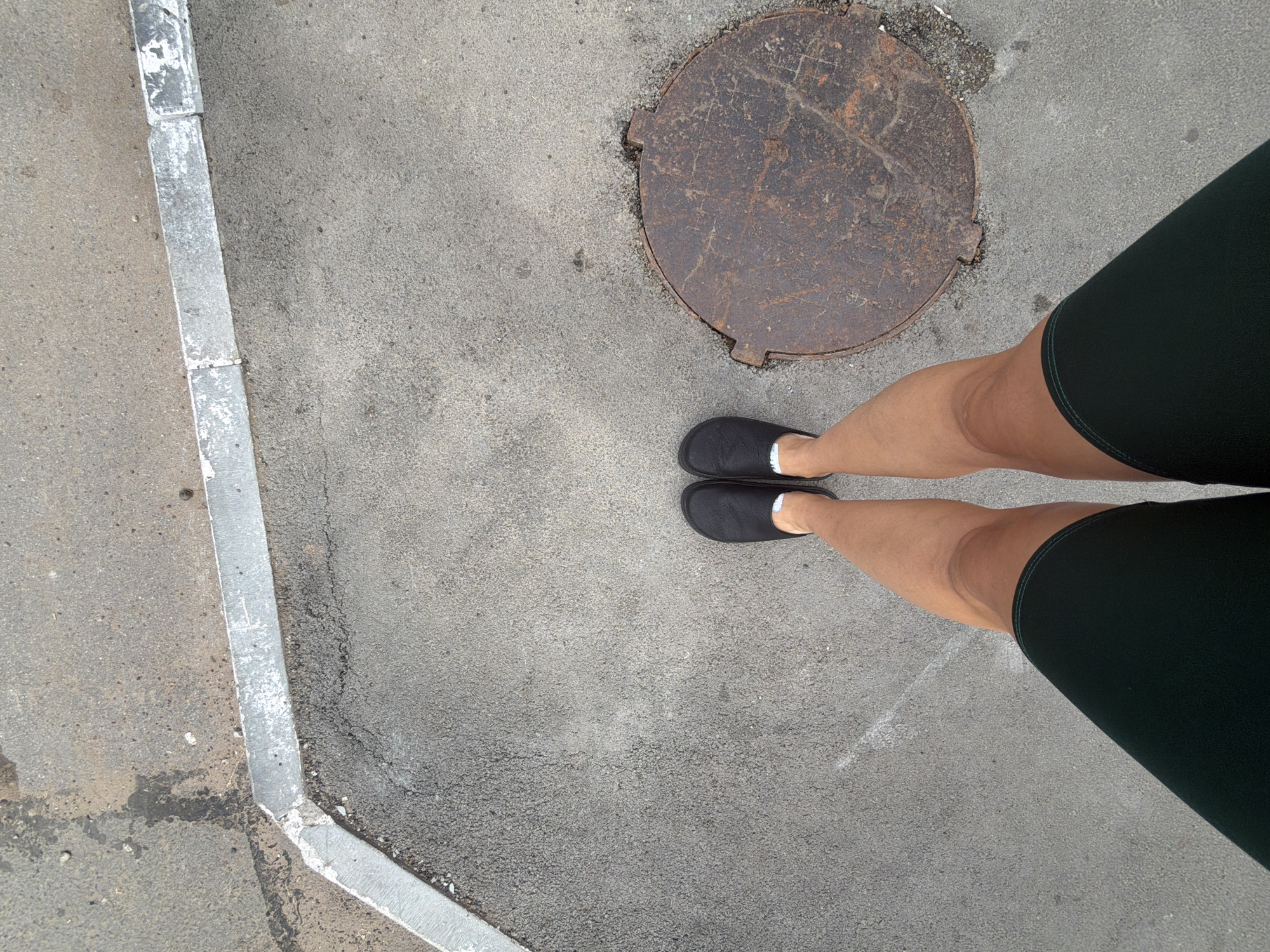 A person wearing black clogs and dark green shorts stands on asphalt. To the left of the person's feet, a concrete curb runs diagonally across the frame, transitioning to a grey sidewalk. To the right, a rusty, circular manhole cover is visible on the asphalt. The scene appears to be an outdoor urban environment, potentially a sidewalk or street in Votkinsk, Russia.