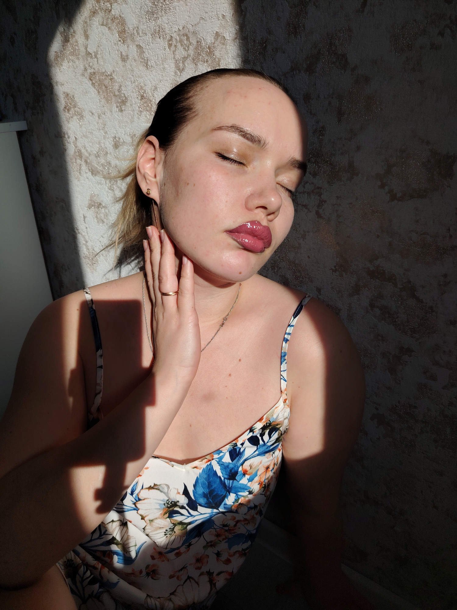 In the city of Марковский, Russia, a female individual is depicted in a medium shot, leaning against a textured wall. She has fair skin, with visible blemishes on her forehead and cheeks. Her eyes are closed, and her lips are full, covered in a glossy, dark pink substance. Her hair, light in color, is pulled back, with some strands visible near her left ear. She wears a small gold stud earring with a dangling gold chain in her left earlobe and a thin gold ring on the fourth finger of her left hand. A delicate gold chain necklace is also visible. She is dressed in a floral-patterned garment with thin straps, featuring white, blue, orange, and green colors. Her left hand is raised, with fingers gently touching her neck and jawline. Strong, directional sunlight illuminates the left side of her face and upper body, casting distinct shadows on the light-colored, patterned wall behind her. A narrow white vertical object is partially visible on the far left edge of the frame.