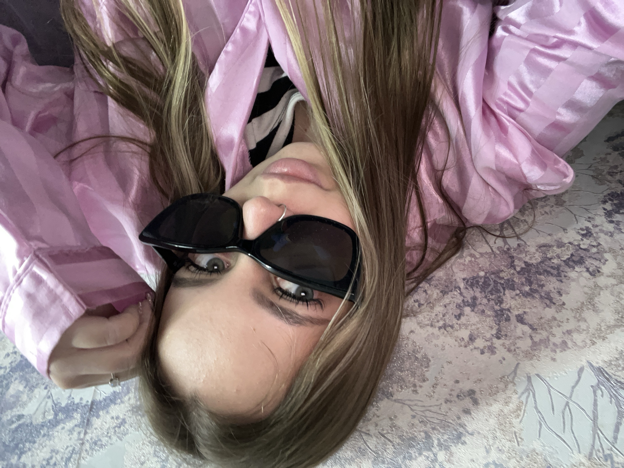 A person with fair skin, light brown hair, and wearing black sunglasses is lying on a surface with a patterned design. The person is also wearing a pink striped pajama top and a striped shirt underneath. The location, Novopokrovskaya, Russia, is not discernible from the image.
