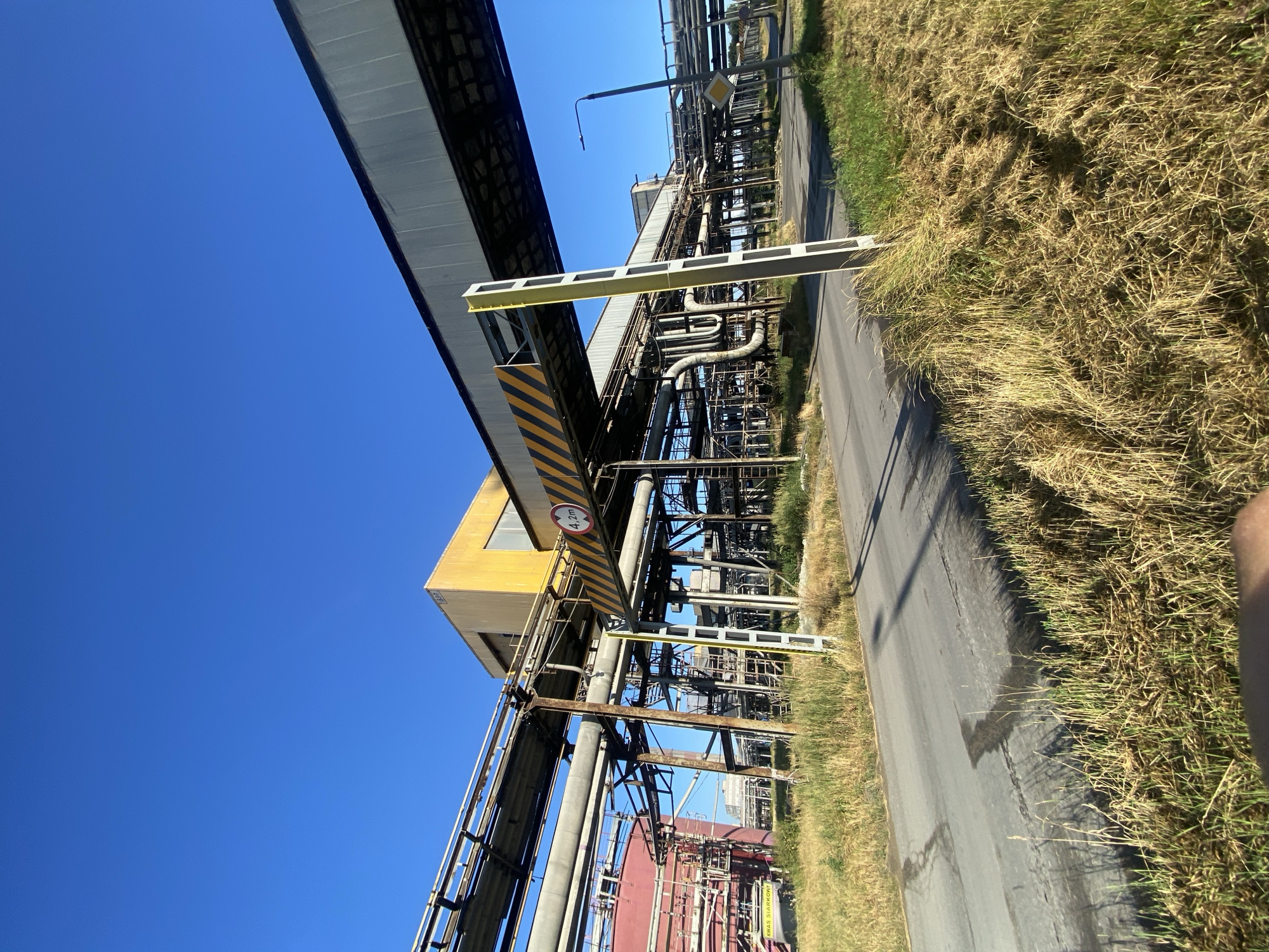 This is a low-angle, wide shot of an industrial complex under a clear blue sky. In the foreground, an asphalt road is visible, bordered by tall, dry grass. A yellow and black striped sign indicating a height limit of 4.2 meters is suspended over the road by a metal structure. To the left, a large, red cylindrical tank with pipes is partially visible. The background is filled with an extensive network of industrial infrastructure, including numerous pipes, metal supports, and elevated walkways. A yellow, rectangular building is situated above and behind the overhead walkway. The scene suggests an active or formerly active industrial site.