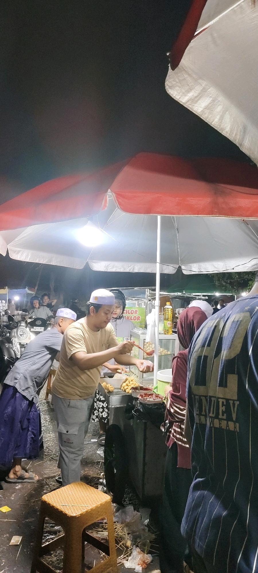 The image depicts an outdoor food stall at night in Martapura, Indonesia. A man wearing a tan t-shirt and a white peci hat is preparing food, holding a ladle over a tray of round, skewered items. To his left, another man in a grey shirt and a blue sarong is leaning over a food cart. Behind them, a woman in a dark hijab is also near the food stall, and another person's back, wearing a dark blue and white striped shirt with "ENDEV WEAR" visible, is in the foreground. The stall is under a large red and white umbrella, illuminated by bright lights. In the background, motorcycles are parked, and other people are visible. The ground is wet and littered with food scraps and plastic wrappers. A small, woven plastic stool is positioned in the lower left of the frame. The overall atmosphere suggests a busy street food environment at night.
