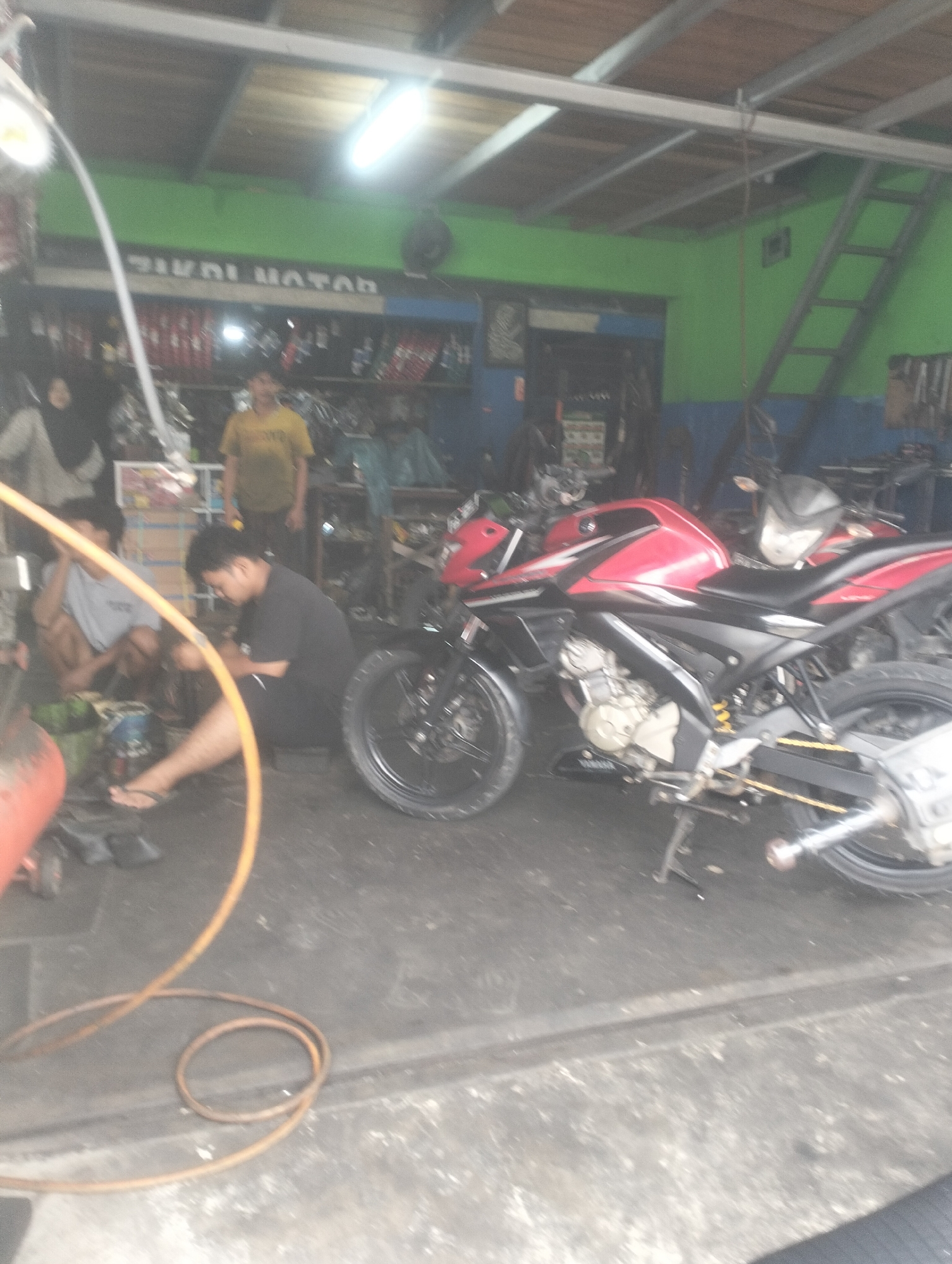 The image depicts a motorcycle repair shop in Dumai City, Indonesia. In the foreground, a red and black motorcycle is parked on a concrete floor. To the left, two individuals are engaged in work, one appearing to be repairing or adjusting something near the motorcycle's wheel, while another sits nearby. Tools and parts are scattered around the work area. In the background, shelves are stocked with various items, possibly motorcycle parts or accessories. The shop appears to be open-air with a corrugated metal roof supported by metal beams. Green and blue paint adorn the walls. A ladder is visible on the right side of the image, leaning against the green wall. A bright fluorescent light illuminates the scene from above.