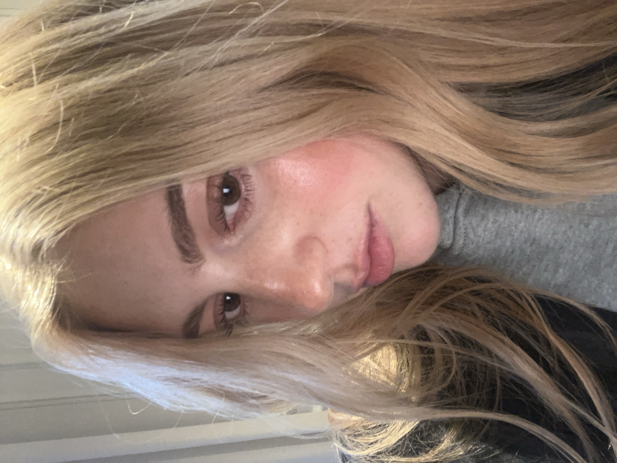 A young adult female with light blonde hair, brown eyes, and fair skin with freckles is depicted in a close-up, angled portrait. She is wearing a grey garment. The location is not identifiable from the image, and there is no indication of Paris, France.