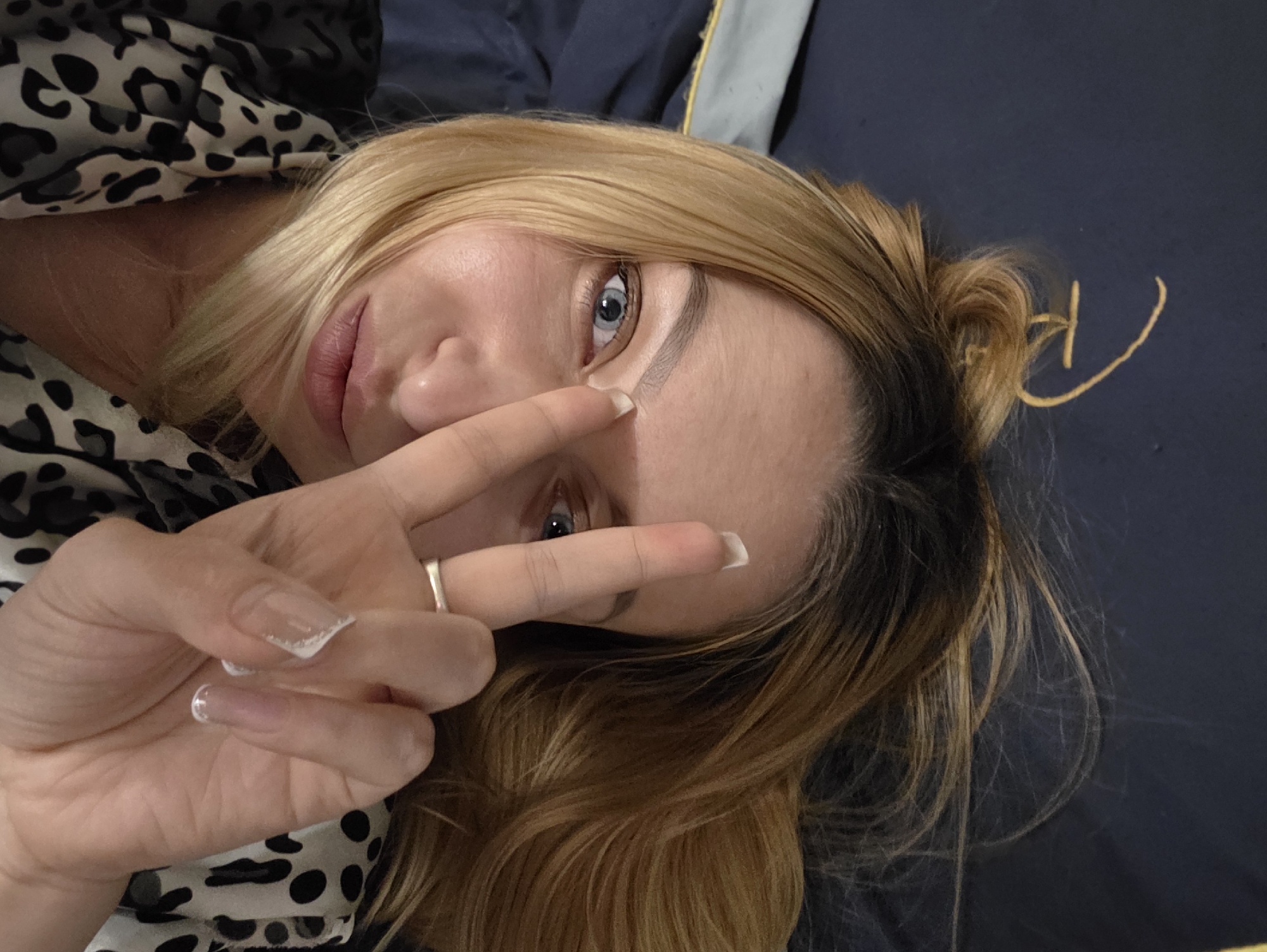 A person, appearing to be female, is depicted lying down, angled towards the camera, with their head resting on dark blue fabric. They possess light brown to blonde hair, styled in a loose bun, with strands falling around their face. The individual has blue eyes and well-defined eyebrows, and their lips are slightly parted. Their right hand is raised, with the index and middle fingers extended in a "V" gesture, partially obscuring the lower part of their left eye. A simple metallic band ring is visible on their right ring finger. Their fingernails are long, clear, and appear to be acrylic or gel, with some featuring a white, possibly french tip, design. The person is wearing a garment with a black and white leopard print pattern. The background primarily consists of the dark blue fabric, likely a bed or pillow. In the upper right corner, a small section of a different dark fabric with a yellow stripe or thread is visible. The scene is illuminated by soft, even lighting, suggesting an indoor setting. There are no identifiable features in the image that would allow for precise geographical location identification within Minsk, Belarus, or any other specific city.
