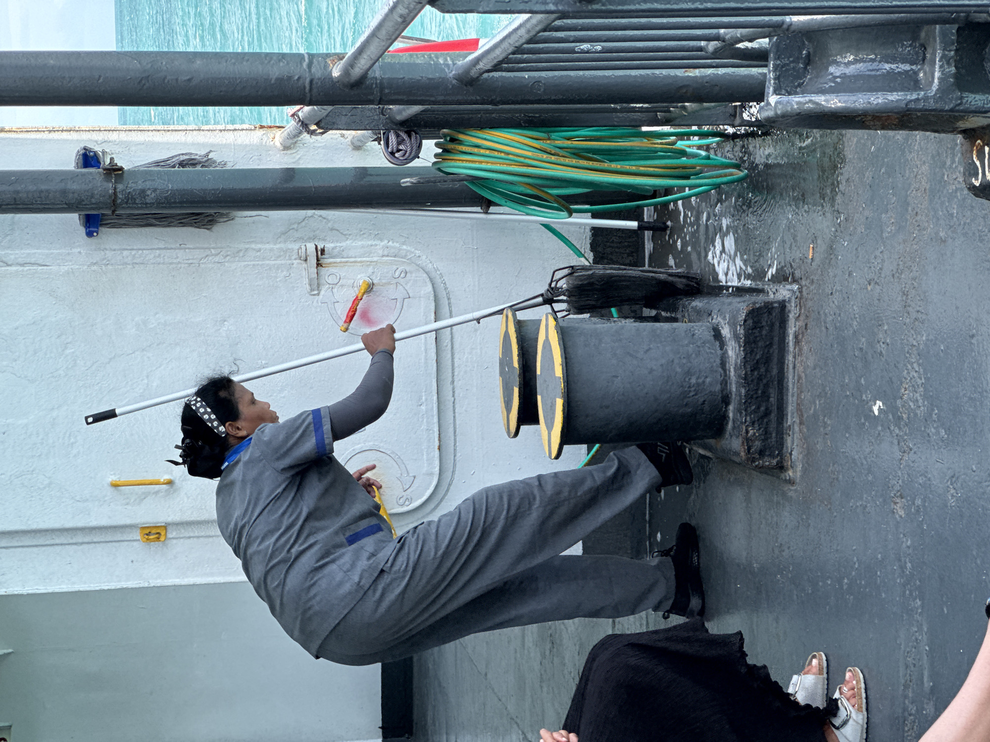 In the city of Unknown, Thailand, a person in a gray uniform with blue trim is mopping the wet deck of a ferry. The person is holding a mop with a dark mop head and a long silver handle. The deck is wet and appears to be soapy, with puddles of water visible. A coiled green and yellow hose is hanging on a vertical post to the right of the person. The background shows the ocean and a white wall with some signage. Several bollards are visible on the deck. There are also other people in the background, some sitting down. The overall scene depicts a cleaning activity on a boat.