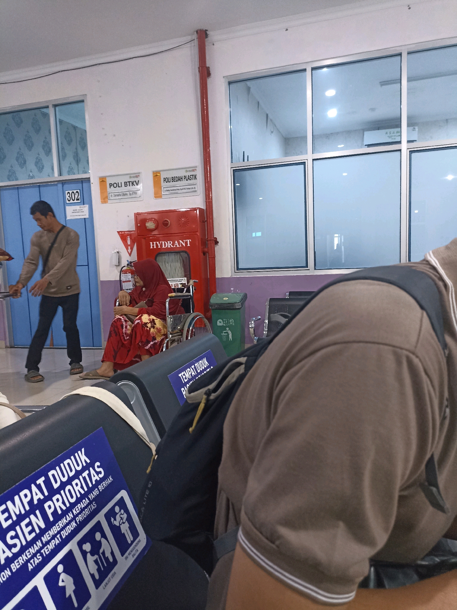 A man in a brown long-sleeved shirt and black pants stands to the left, facing away from the camera. A woman in a red hijab and a red patterned dress is seated in a wheelchair to his right, looking to the left. A red fire hydrant with the word "HYDRANT" is visible behind her. A green trash can is next to the hydrant. In the foreground, a blue sign on a black seat reads "TEMPAT DUDUK PASIEN PRIORITAS" (Priority Patient Seating). The scene appears to be a waiting area, possibly in a hospital or clinic, with large windows in the background. The room is brightly lit, and the walls are white. The overall impression is one of a public waiting space with people and essential services.