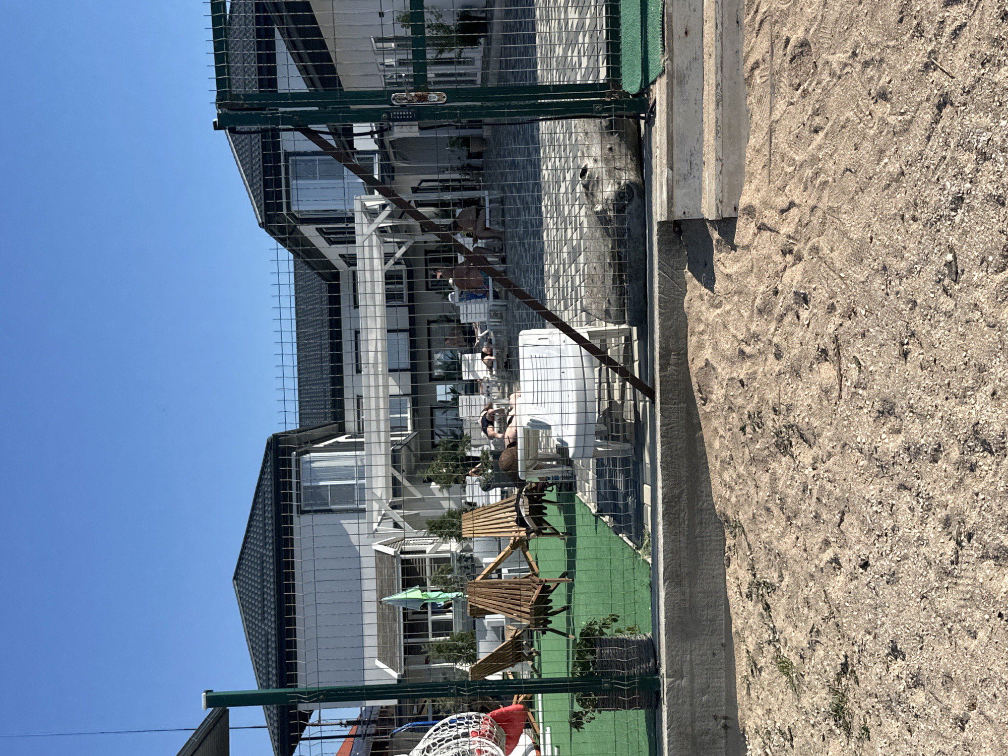 In Golubitskaya, Russia, a sun-drenched courtyard is visible behind a wire fence, featuring a modern, white building with a dark roof. Several people are relaxing on lounge chairs on a paved area. In the foreground, a sandy area with scattered shells and small plants transitions to a bright green artificial turf. Wooden chairs and a green umbrella are set up on the turf. The scene appears to be a resort or hotel courtyard on a clear, sunny day.