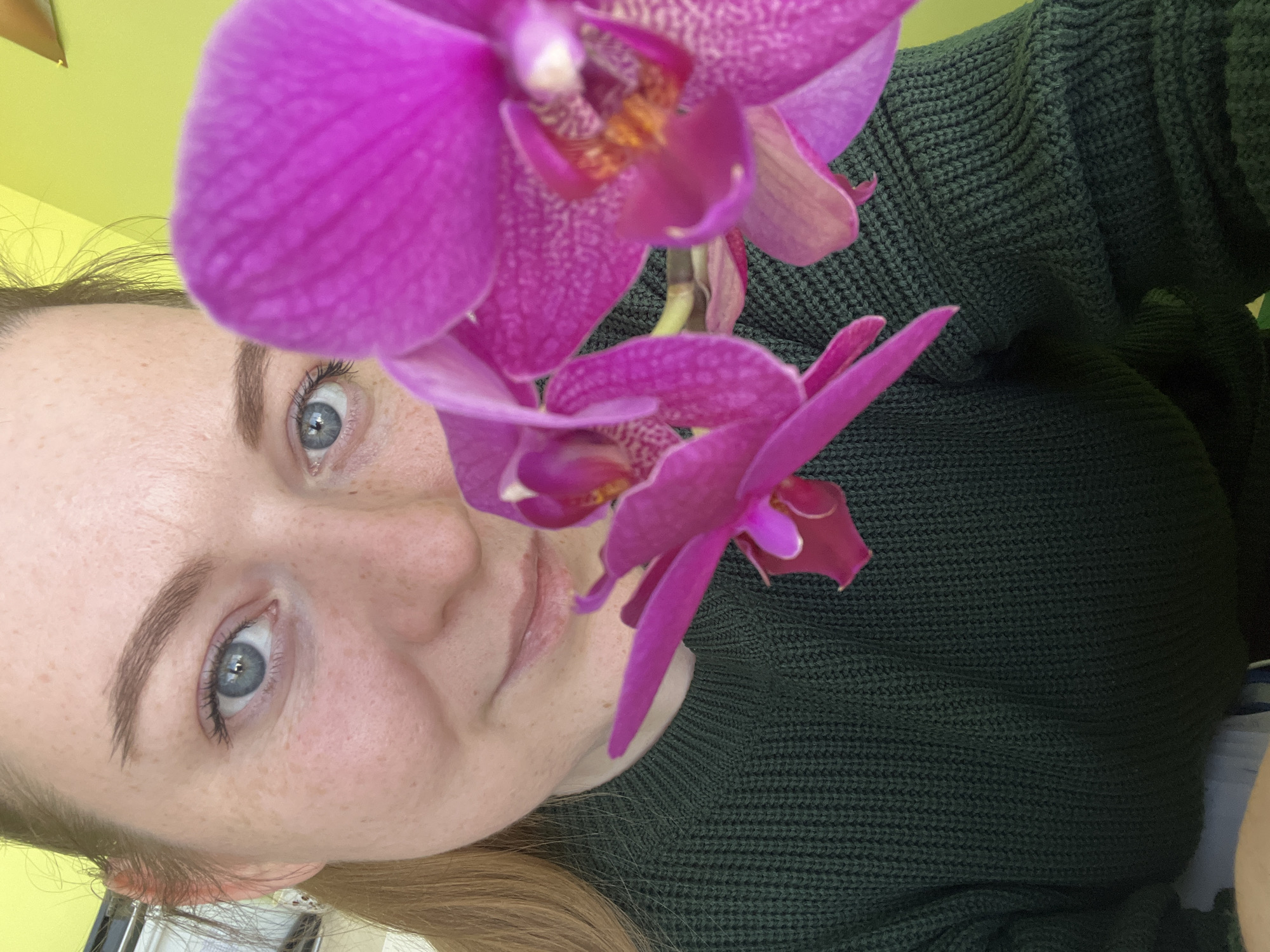 A woman with fair skin, freckles, and light blue eyes looks directly at the camera, framed by two vibrant pink orchids. She is wearing a dark green knit sweater. The background includes a yellow wall and glimpses of blonde hair. The scene takes place indoors. The location, Klin, Russia is not discernible in the image.