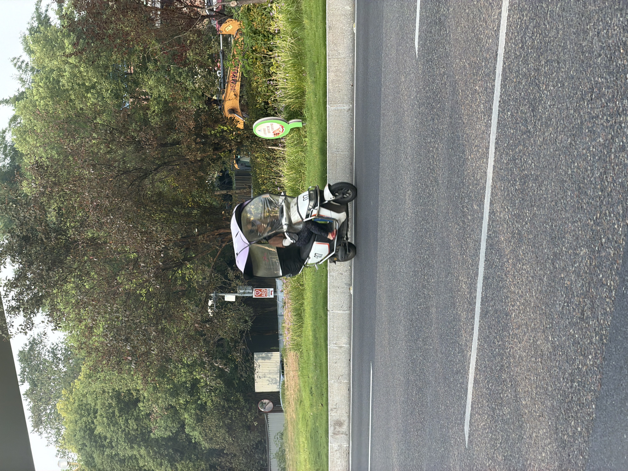 A person is riding a white motorized scooter equipped with a purple umbrella canopy, a transparent enclosure around the handlebars and front wheel, and a rear storage box. The scooter has the number "07" displayed on its side. The rider is wearing dark pants and red shoes. The scooter is on a paved road with white lane markings, next to a concrete curb and a grassy area. In the background, there are trees, a building with a corrugated metal exterior, a traffic sign with an AED symbol, a yellow construction vehicle, and a green circular sign with Chinese text and an image of a bird. The scene appears to be in a city setting in Jiangjin, China.