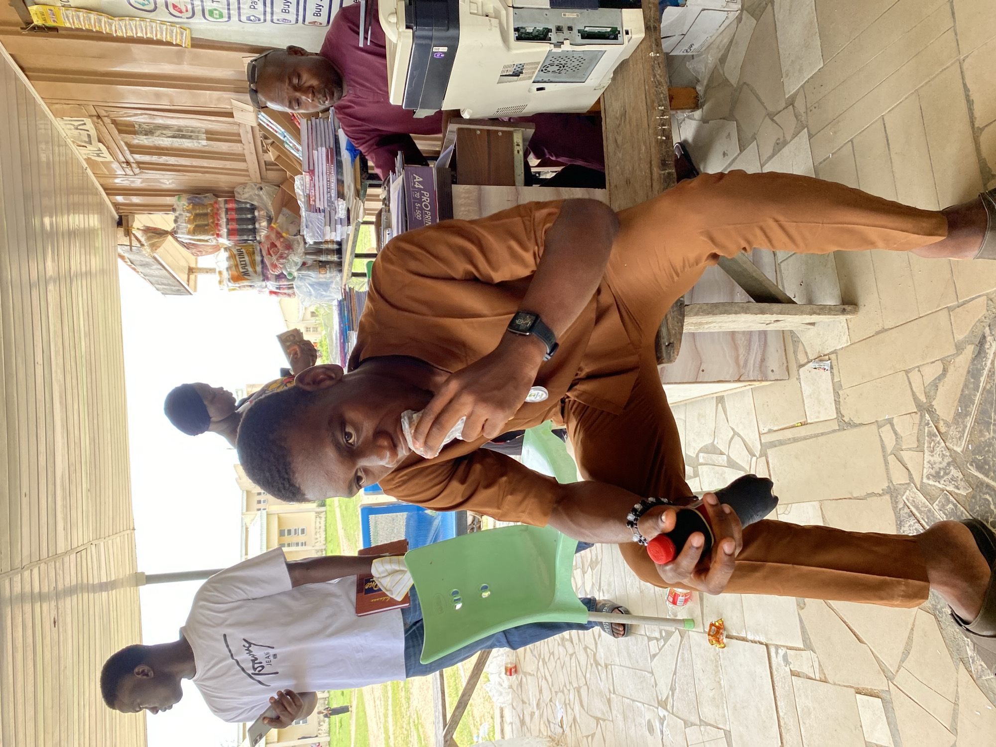 The image depicts an outdoor scene at a roadside kiosk in Yali, Nigeria. In the foreground, a young man wearing a brown outfit is seated on a wooden bench, eating from a white wrapper and holding a black object with a red top in his left hand. He wears a wristwatch on his right wrist. To his left, a green plastic chair is visible. Behind him, another young man in a white t-shirt stands holding a mobile phone and a book. Further in the background, a woman in a dark headwrap is partially visible. To the right of the seated man, an older man in a maroon garment is behind a counter, near a printer. The kiosk displays various items, including bottled drinks, snacks, and stacks of notebooks. The ground is paved with irregular tiles. The environment suggests a commercial area within Yali, Nigeria.