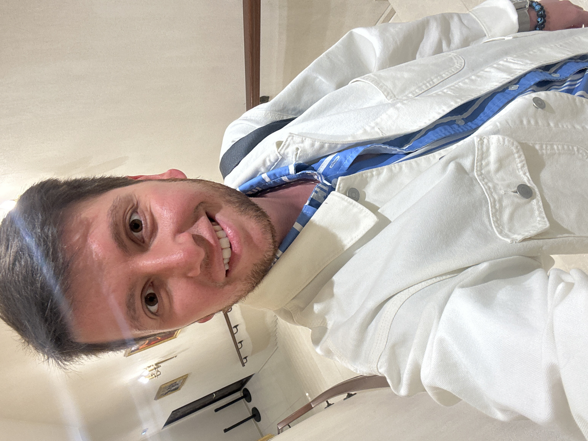 The media file is a close-up, low-angle, portrait-style selfie of a young man with short brown hair and a beard. He is wearing a white denim jacket over a blue and white striped shirt. His eyes are open, and he is smiling broadly, showing his teeth. He is wearing a watch on his left wrist with dark blue beads and a metallic band. The background appears to be an indoor setting, possibly a staircase or hallway, with light-colored walls and some framed artwork or decorative items on the wall. The lighting suggests it could be daytime or well-lit indoors. There is no visible text in the image. The overall mood conveyed is cheerful and casual.