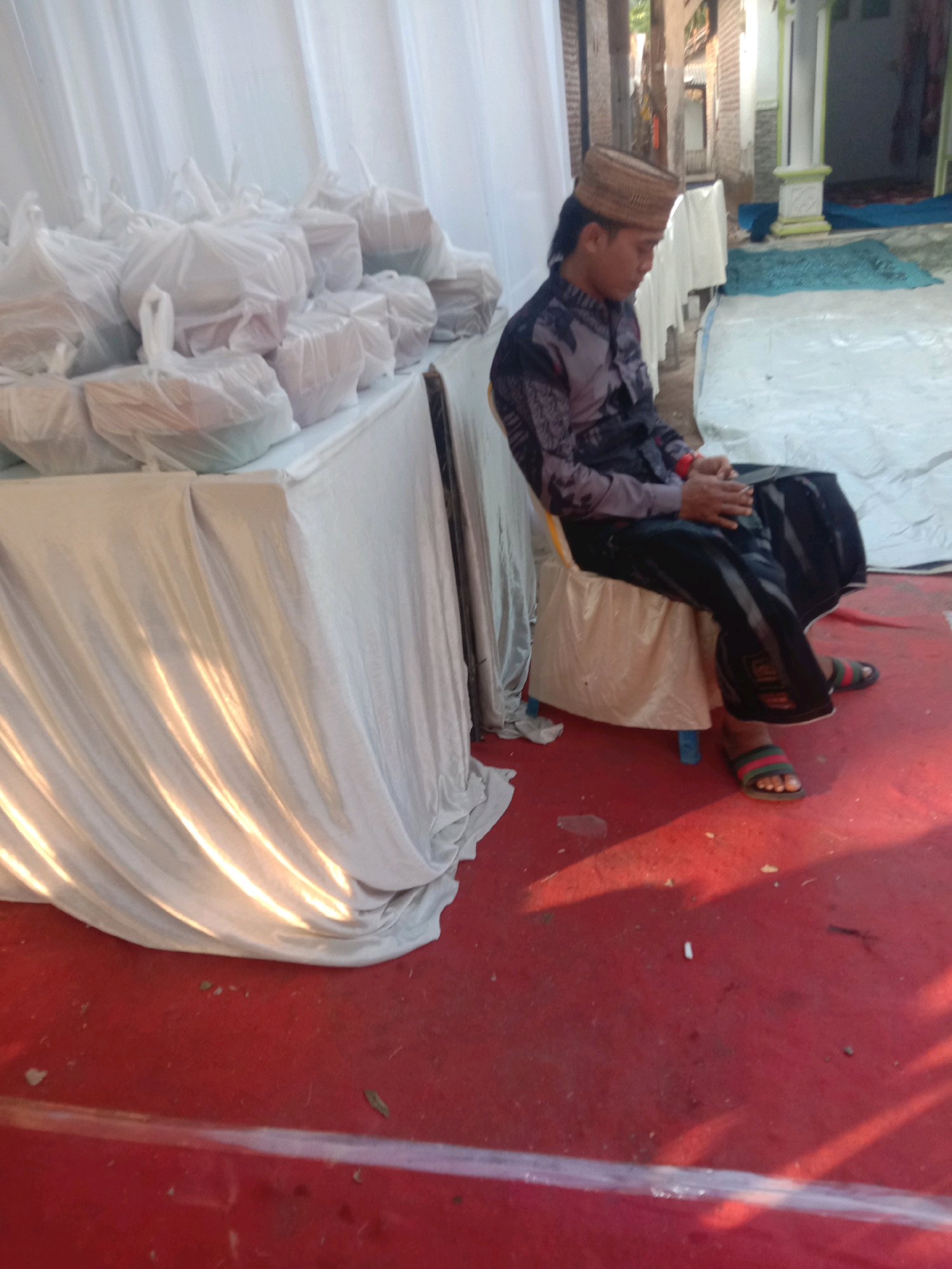 The image depicts an outdoor scene in Karang Anyar, Indonesia, likely during an event or ceremony, with a red carpet laid out on the ground. A young man, dressed in traditional attire including a patterned shirt, dark trousers, and a woven cap, is seated on a cushioned chair. He is looking down at his hands, which are clasped in his lap. To the left of the frame, a table is draped with a white tablecloth, laden with numerous white plastic-wrapped packages. The background shows a white tent or canopy, and hints of buildings and foliage. The lighting suggests it is daytime, with sunlight casting shadows and highlights on the white fabric and the red carpet.