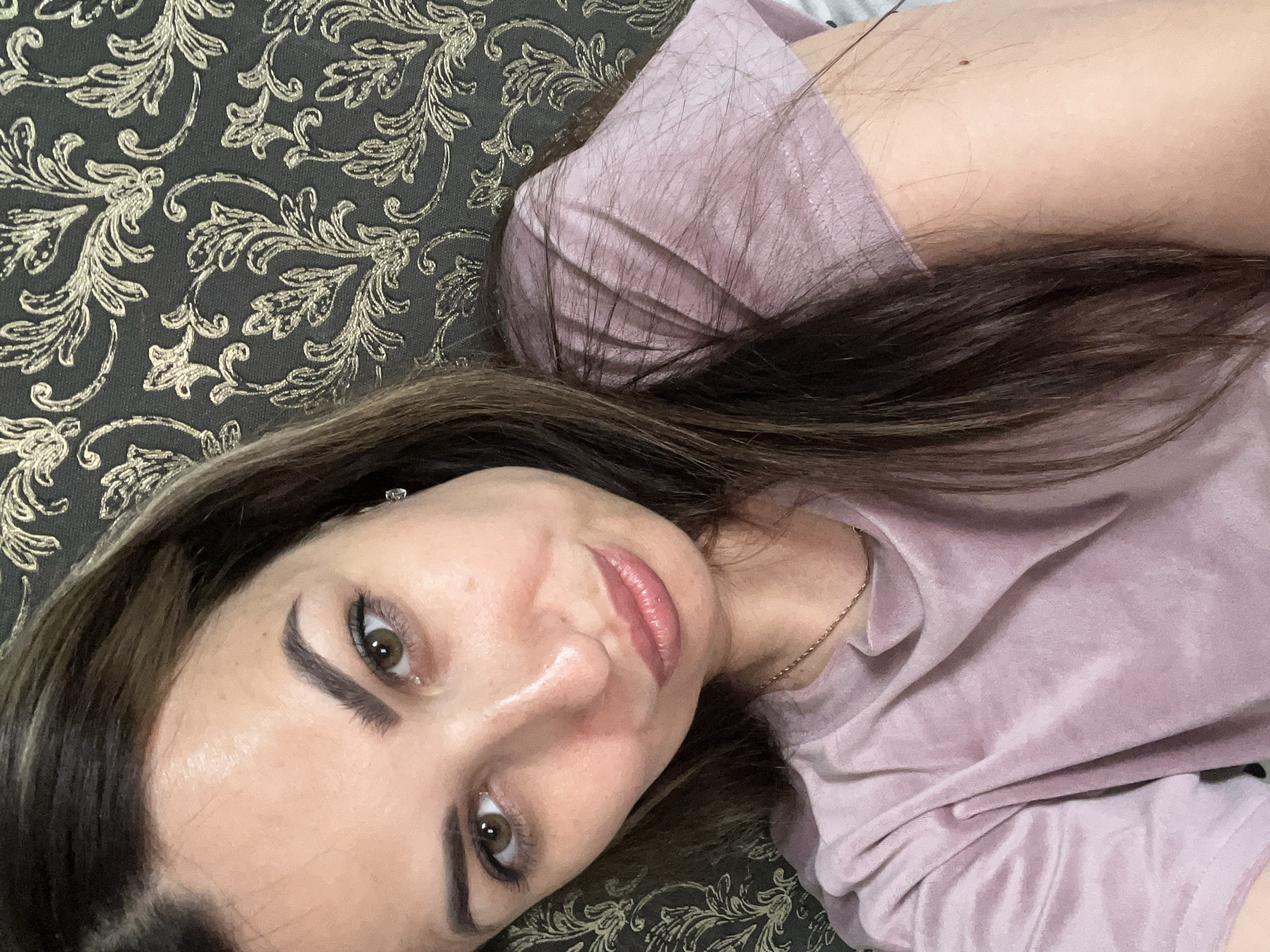 A woman with brown hair and brown eyes is taking a selfie. She is wearing a light purple t-shirt and a gold chain necklace. She has her mouth slightly pursed and is looking towards the camera. The background is a dark fabric with a gold damask pattern. The lighting is soft and the image has a slight blur. The scene is indoors.