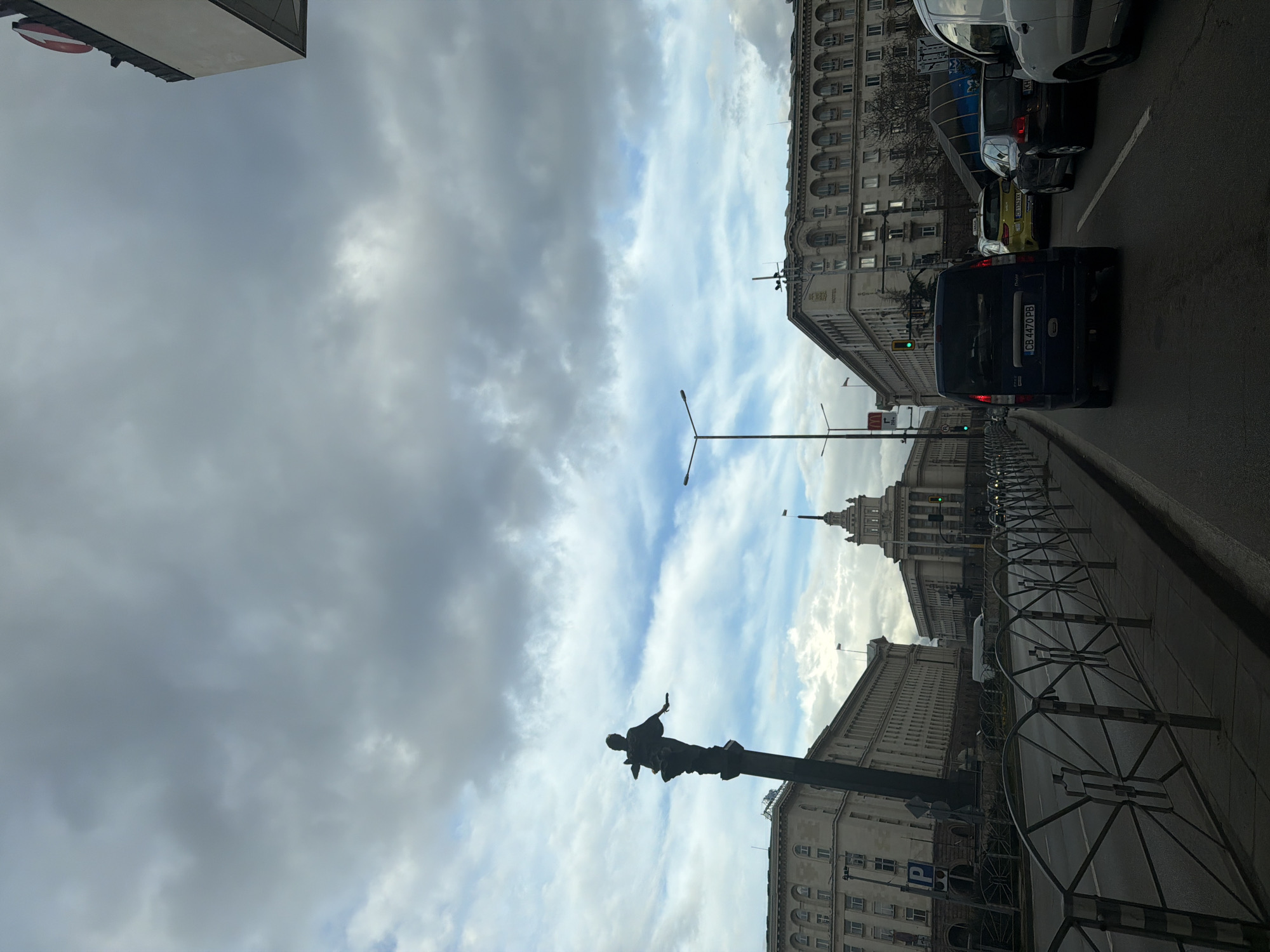 The image shows a street view in Sofia, Bulgaria. On the right side of the frame, there are several cars, including a dark blue van with the license plate "CB 4470 PB." The background consists of buildings and a cloudy sky. A statue is situated on a pedestal on the left side of the frame. A building with a pointed tower can be seen in the distance. A light post with two horizontal crossbars is near the center of the image. A row of circular, metal barriers are on the right sidewalk along the buildings.
