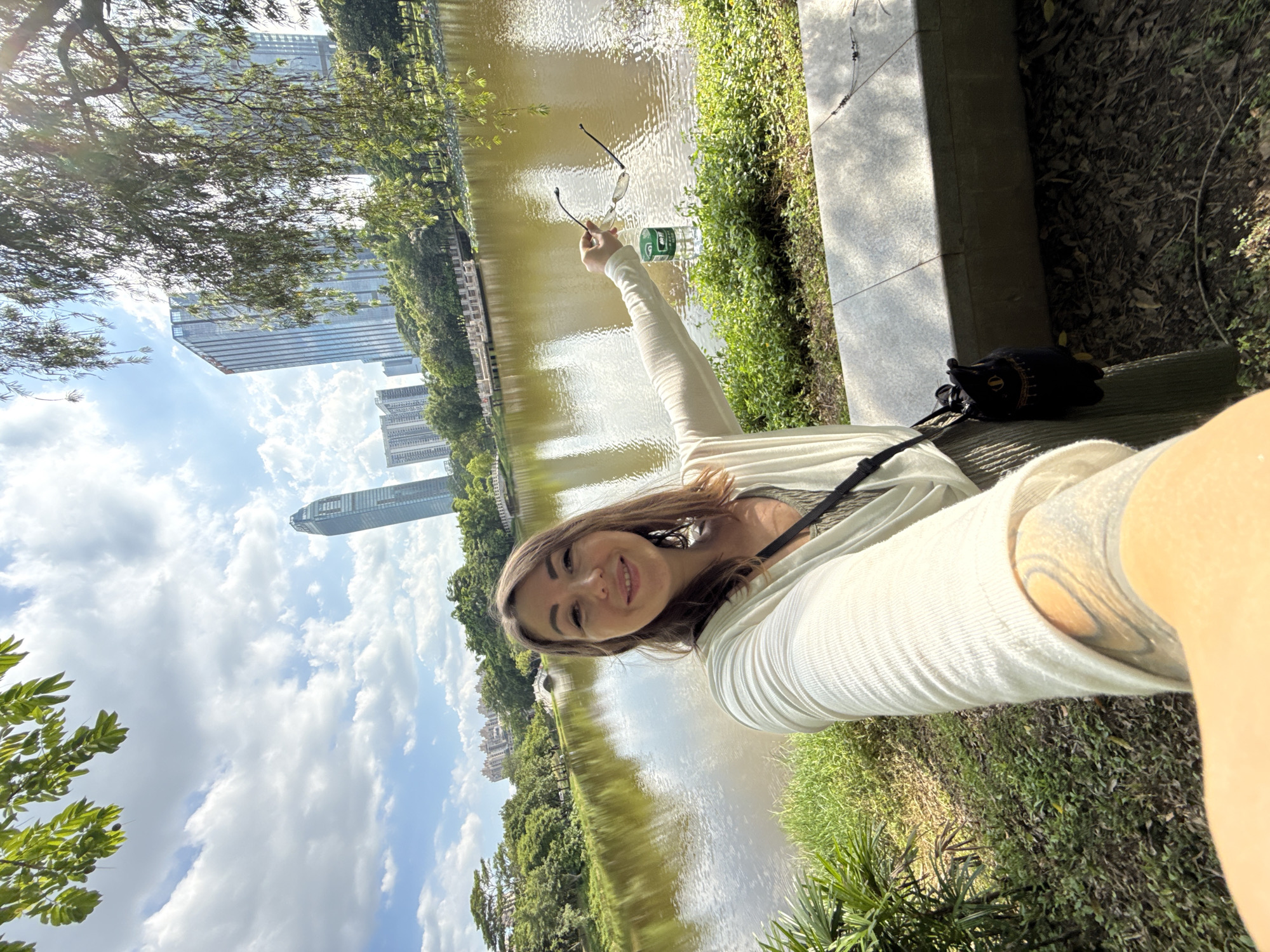 A woman takes a selfie in front of a lake in Huizhou, China. She is wearing a white cardigan over a patterned top and khaki pants. She holds a pair of sunglasses and a bottle of water in her right hand. Behind her, the lake reflects the blue sky and clouds, as well as trees and modern buildings. The scene appears to be a park, with grass, trees, and a stone bench.
