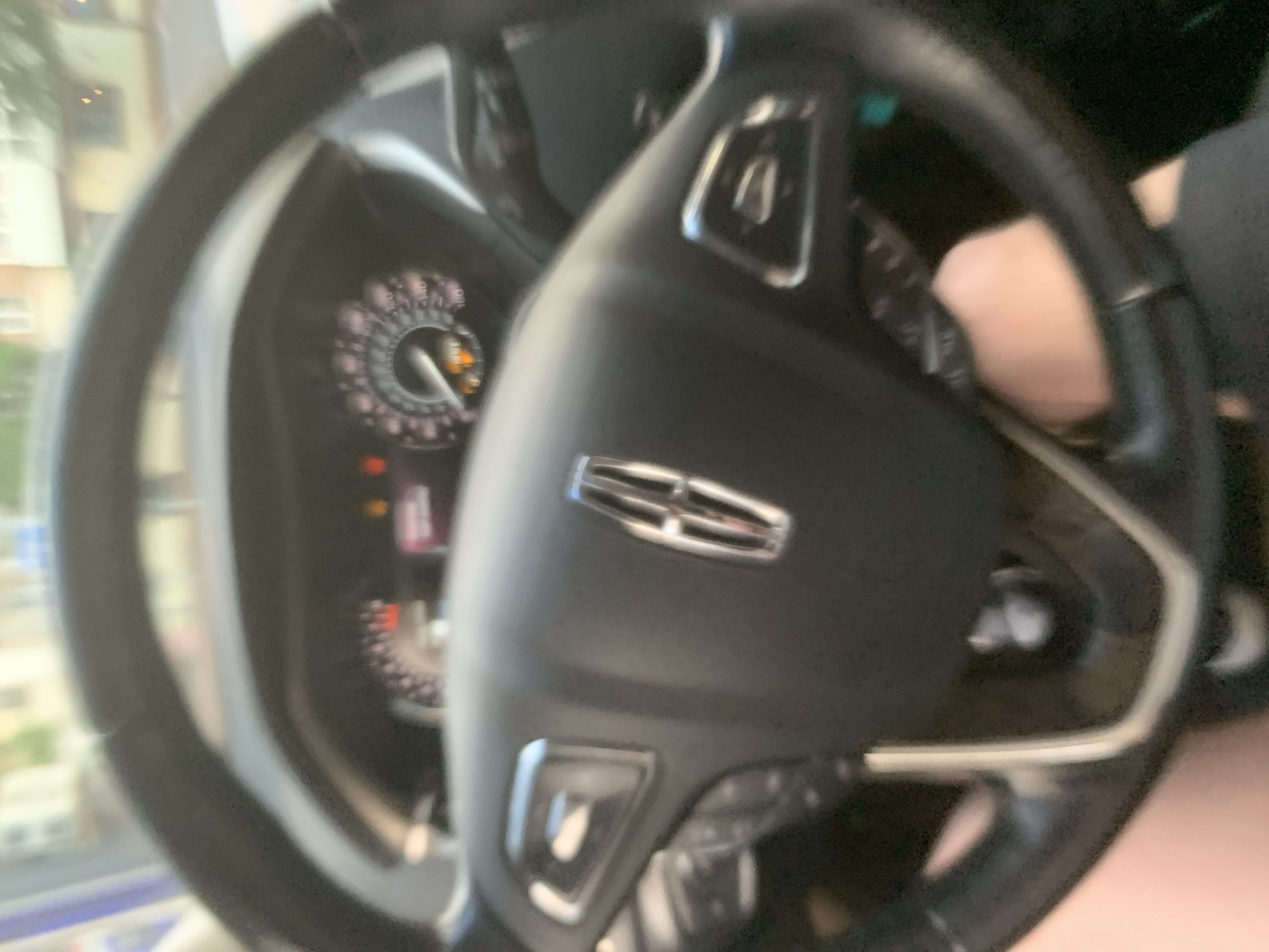 The image shows the interior of a car, focusing on the steering wheel and dashboard. The steering wheel is black and appears to be made of leather. In the center of the steering wheel is a silver emblem with horizontal lines, which appears to be a car logo. To the left of the steering wheel, a portion of the car's instrument cluster is visible, displaying a circular gauge with orange and white markings, along with some illuminated indicator lights. A person's hand is partially visible on the right side of the steering wheel, suggesting the car is being driven or is about to be driven. The background is blurred but hints at an outdoor scene with buildings and greenery, possibly indicating a daytime setting. The overall lighting suggests it is not dark, though no specific time of day can be definitively determined. There is no visible text in the image.
