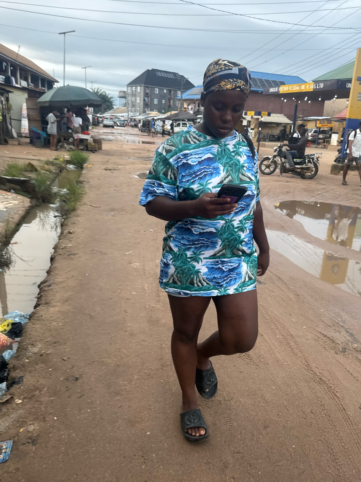 A young Black woman with a patterned headscarf and a blue and green tropical-print t-shirt dress walks on a dirt road in Ihiagwa, Nigeria. She is holding a smartphone in her right hand and looking down at it. She wears black slides. The road is lined with shops, some with awnings, and a canal with standing water and debris on the left. In the background, a man rides a motorcycle, and other pedestrians and vehicles are visible. The sky is overcast.