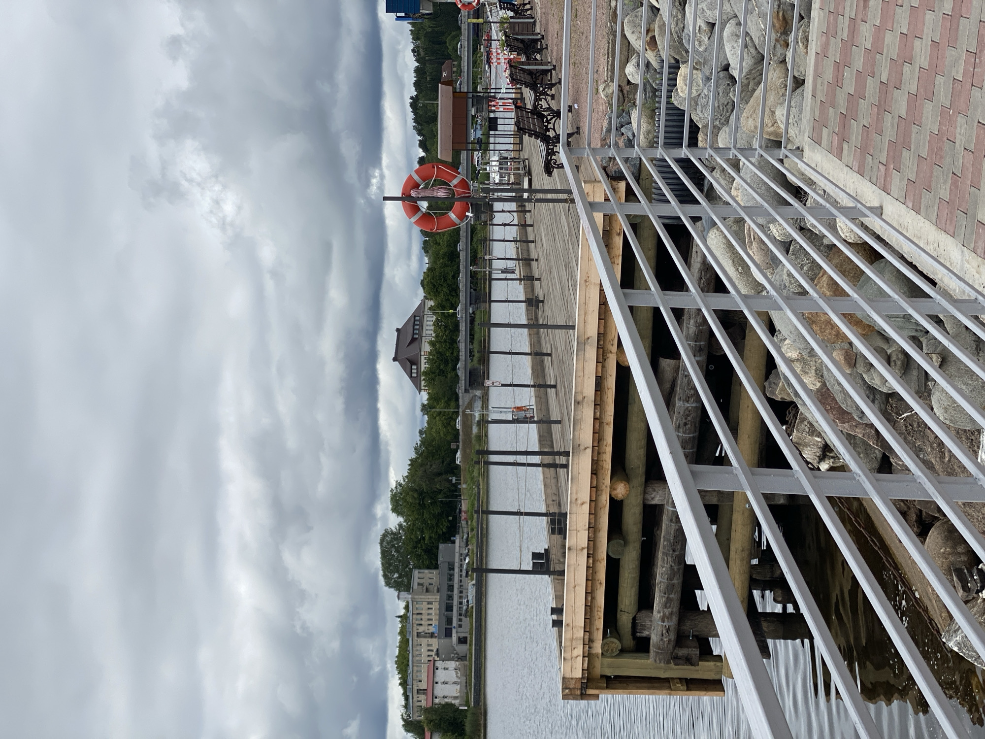 The image shows a wooden pier extending into a body of water under a cloudy sky. The pier has a metal railing along its edge, and several mooring posts are visible. In the background, across the water, there are buildings and trees. A bridge can be seen in the distance. A red life preserver is hanging on a post on the pier. Benches are also present along the waterfront. The scene appears to be a public area, possibly a promenade or park, in the city of Sortavala, Russia. The water is calm, and the overall atmosphere is somewhat subdued due to the overcast weather. The foreground features a paved walkway with red and grey brickwork.