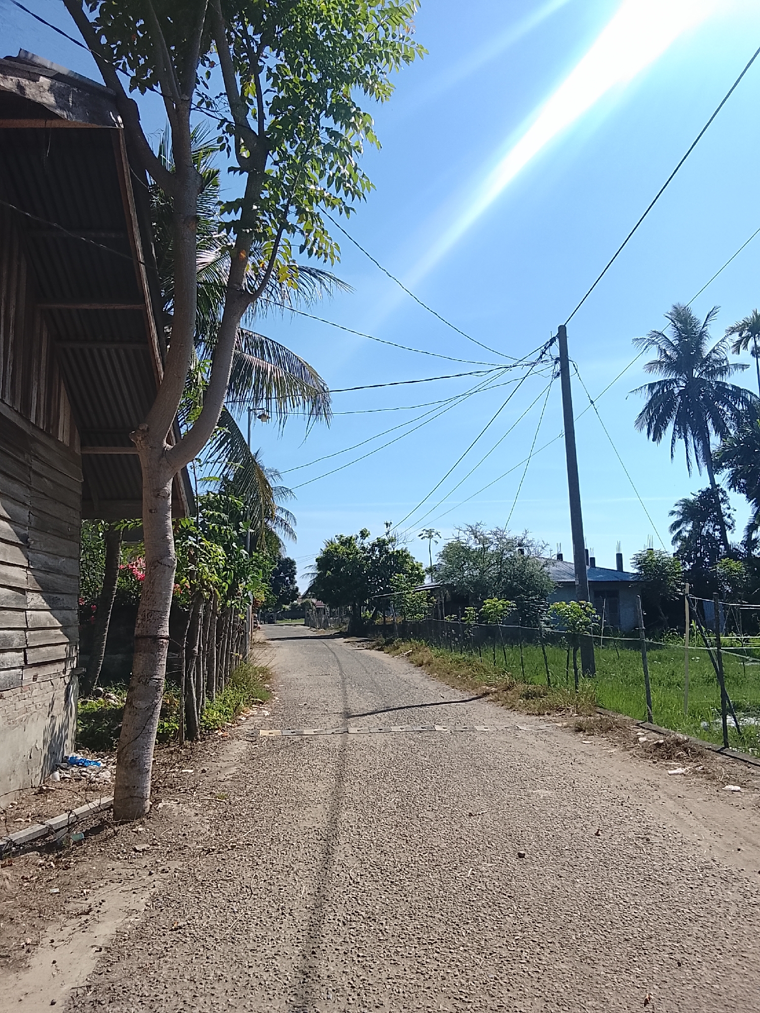 A rural dirt road stretches into the distance under a bright blue sky, with the sun glaring in the upper right quadrant, creating a strong lens flare. To the left, a weathered wooden structure with a corrugated metal roof stands adjacent to the road. A young tree with light-colored bark is positioned in front of this structure. Beyond the tree, a line of thin wooden posts supports a fence that runs along the side of the road. Lush green foliage and palm trees are visible on both sides of the road, interspersed with small houses and buildings. A utility pole with a complex network of wires stands near the center of the image, on the right side of the road. The ground on the road is covered in small gravel and dirt, with some scattered debris near the base of the wooden structure. The scene appears to be a snapshot of daily life in a village, possibly in Geuleudah, Indonesia, given the tropical vegetation and building styles.