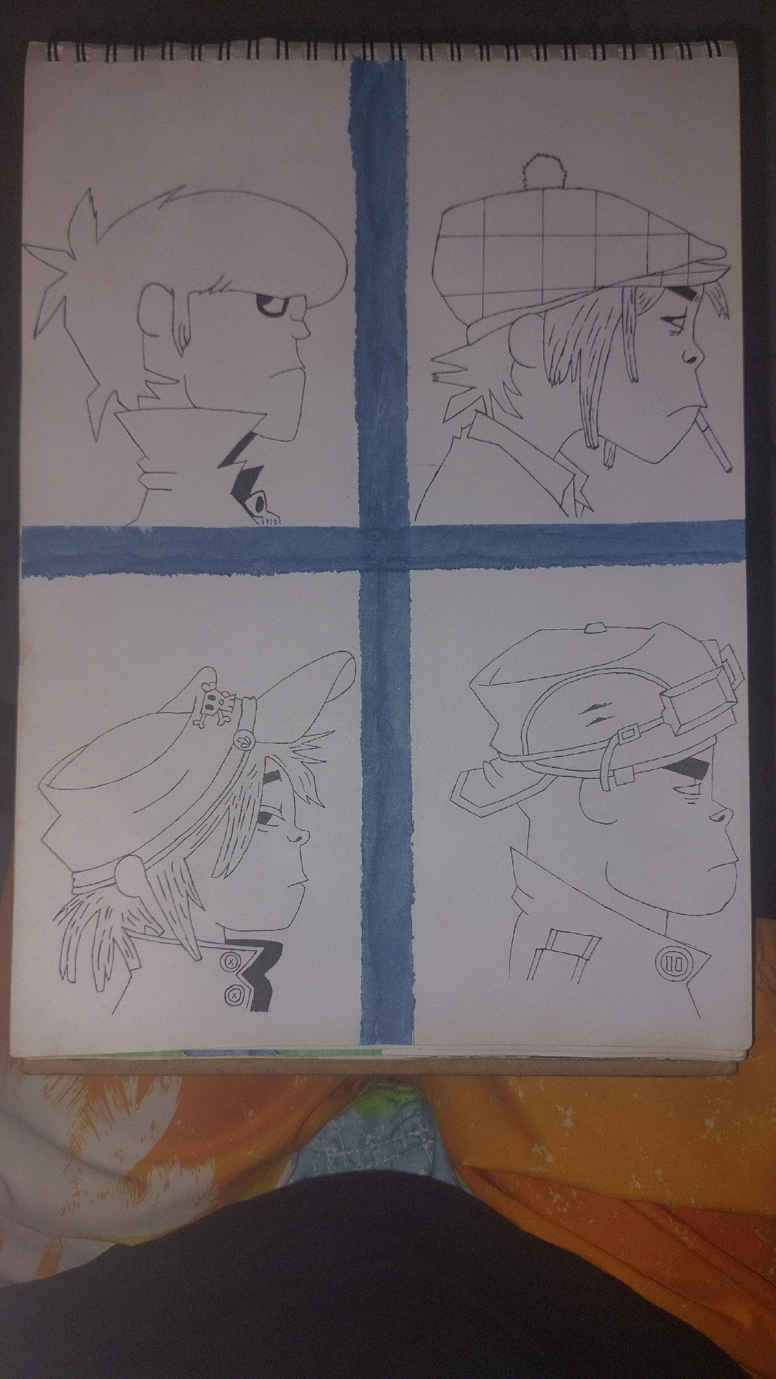 The image is a drawing on a spiral-bound notebook, divided into four panels by blue lines. Each panel features a character from the Gorillaz band, drawn in profile facing right. The top left panel shows 2-D with spiky hair and a stern expression. The top right panel shows Murdoc Niccals wearing a flat cap and smoking a cigarette. The bottom left panel depicts Noodle with a hat adorned with a skull and crossbones. The bottom right panel shows Russel Hobbs wearing a cap with goggles. The notebook is lying on a colorful, patterned fabric. The image does not contain any information about the city of Nairobi, Kenya, or any specific geographic location.