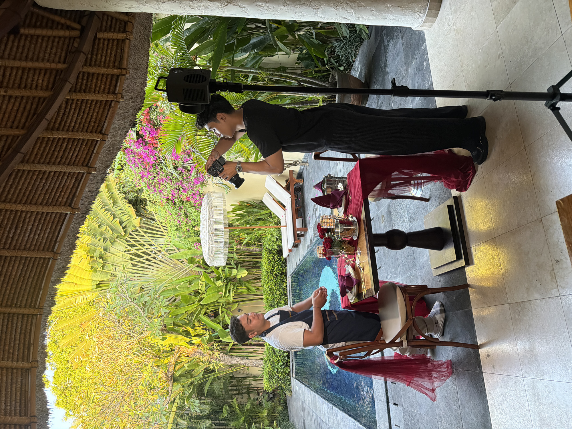 Two young men are in an outdoor setting, likely a resort or a private villa, with a swimming pool and lush tropical greenery in the background. The man on the left, wearing a white t-shirt and a dark apron, stands beside a dining table set for two with red tablecloths, roses, and champagne glasses. He appears to be preparing or arranging something on the table. The man on the right, dressed in a black t-shirt and dark pants, is holding a camera and taking a picture of the table setting. A professional lighting setup is positioned behind him. The scene is set under a thatched-roof structure, suggesting an open-air dining or event area. The overall atmosphere suggests a romantic or upscale dining experience being documented.