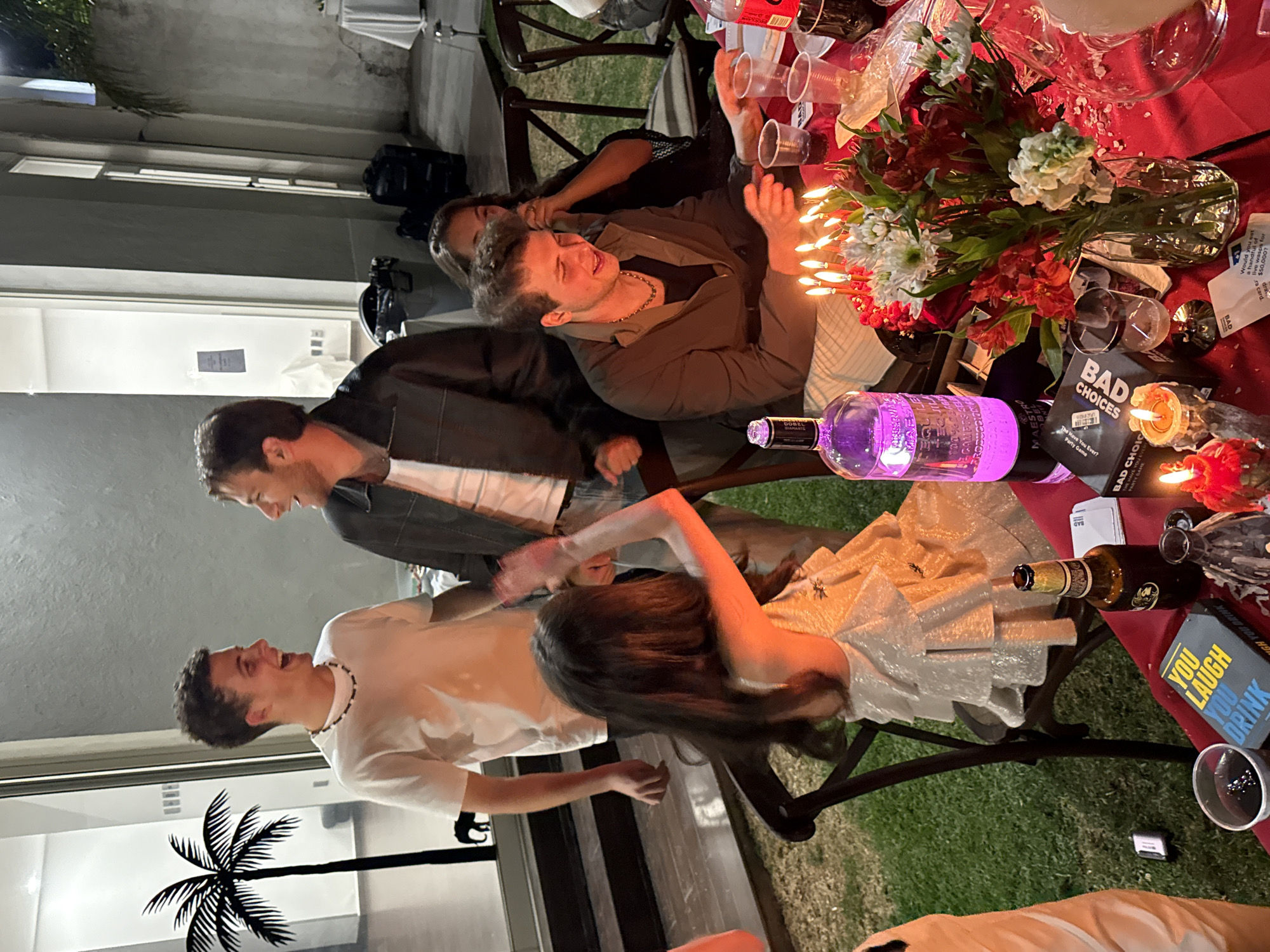The image shows a group of people gathered around a table outdoors, likely at night, judging by the lighting and the presence of candles on the table. There are four people partially visible at the table. The tabletop is red and covered with various items including a floral centerpiece in a glass vase, a bottle of alcohol with a purple illuminated body, a card game box labeled "BAD CHOICES", a bottle of beer, drinking glasses, and other miscellaneous items. One person at the table is wearing a dress with shimmering details, and another is wearing a white shirt with a beaded necklace. Additional individuals are partially visible, including one wearing a brown jacket and another wearing a dark shirt. A decorative metal palm tree is visible outside a window. The location appears to be Huixquilucan de Degollado, Mexico, based on OCR content that may be related to local context.