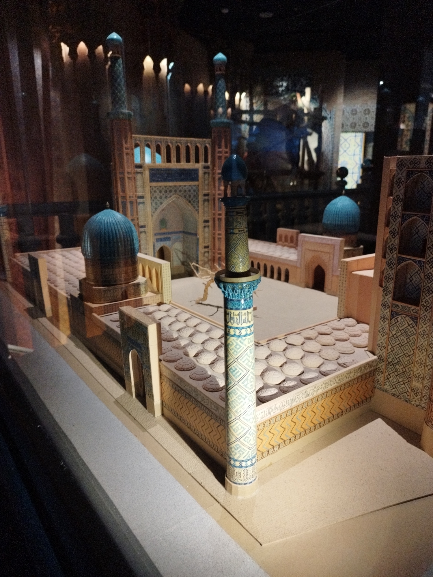 This is a photograph of a miniature architectural model of a mosque. The model is displayed in a glass enclosure, possibly in a museum. The mosque features a central courtyard surrounded by buildings with intricate blue and yellow geometric tilework. Several minarets with blue domes rise from the structure. The courtyard floor is tiled with numerous circular elements. In the background, blurred figures and architectural elements suggest a larger exhibition space. The lighting is dim, with spotlights illuminating the model. The photograph is taken from a slightly elevated, angled perspective, looking down and across the model.