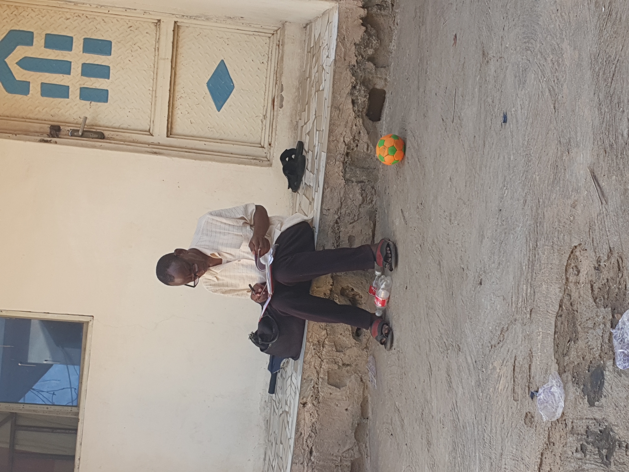 The image shows a man in Lokoja, Nigeria, sitting in a narrow space between a building and a concrete walkway. The man is wearing glasses, a light-colored, short-sleeved shirt, and dark pants with sandals. He is bent over, writing on a pad of paper with a pen. A bag and a blue jacket are on the ground near him. Two empty plastic Coca-Cola bottles are also near his feet. In the background is a cream-colored metal door with geometric blue designs. Next to the door, a black object and a green and orange ball are on the walkway. The concrete walkway is textured and shows signs of wear, including litter.