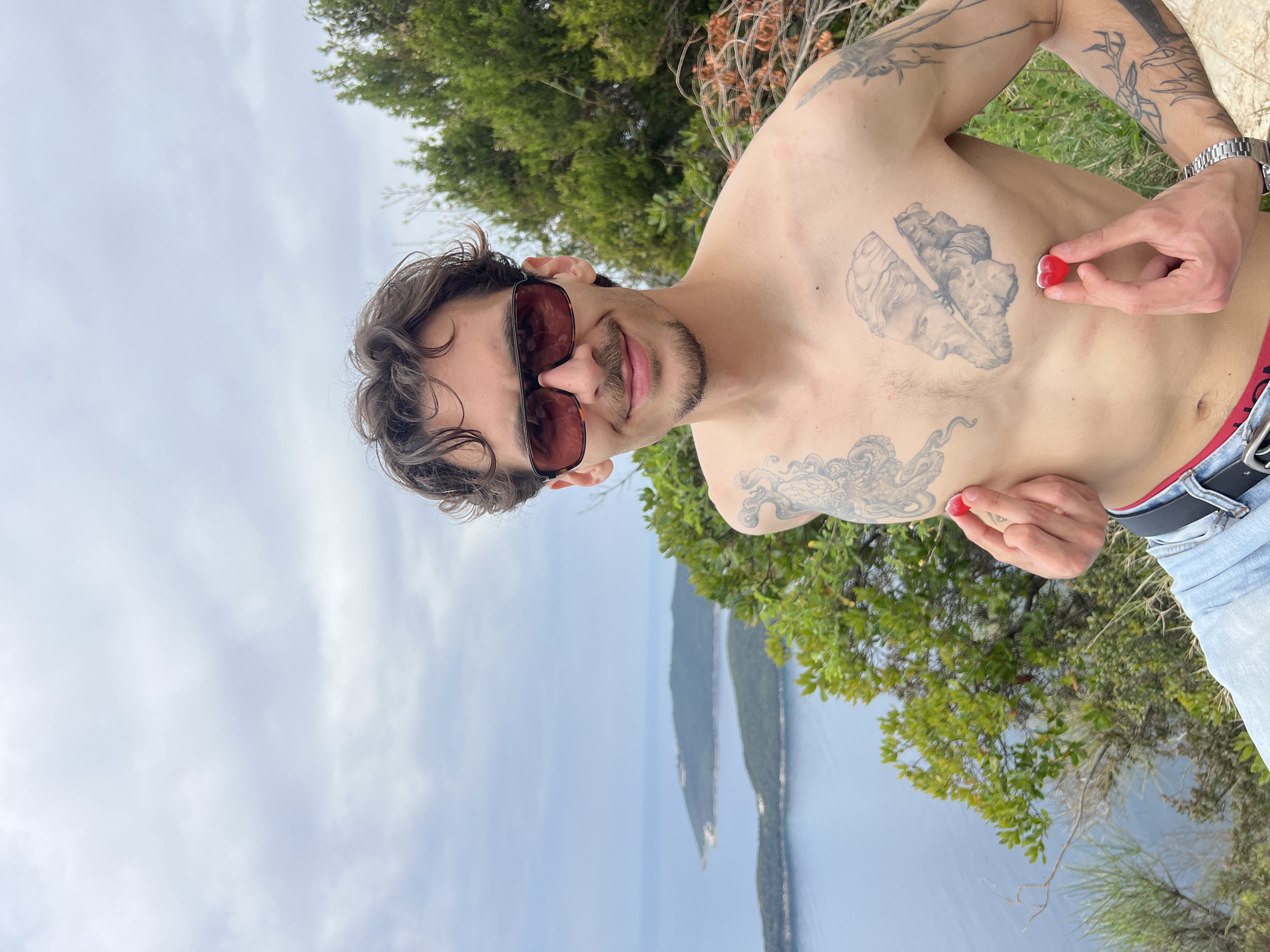 A man is positioned in front of a scenic overlook in Budva, Montenegro. He is shirtless, wearing sunglasses, and holding two red heart-shaped candies. The man has tattoos on his chest and arms. He is also wearing jeans and a watch. Behind him, the Adriatic Sea is visible, along with an island. The sky is partly cloudy. There are trees and shrubs around the man and along the cliffs overlooking the sea.