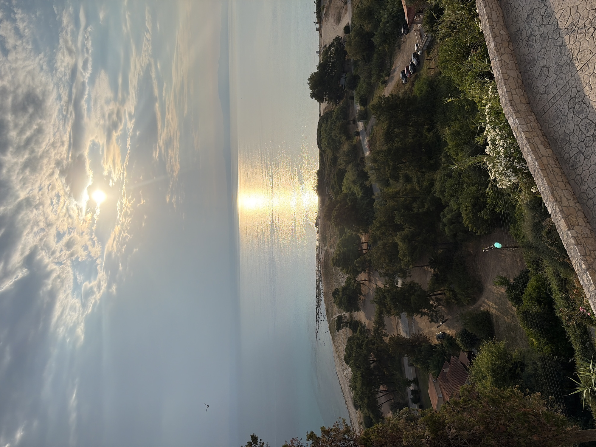 A wide-angle, high-angle shot captures a serene coastal scene at sunset in Kassandra Municipal Unit, Greece. The sun, partially obscured by clouds, casts a brilliant, shimmering reflection across the calm, turquoise sea. A thin strip of sandy beach with scattered rocks separates the water from a dense green landscape.
The foreground is dominated by lush green trees and bushes, with a pathway paved with irregular stone slabs curving into the lower right corner. A stone wall runs along the edge of this pathway. A few cars are parked on a dirt road that winds through the trees. A small cluster of houses with terracotta roofs is visible nestled among the foliage on the left side of the frame. A solitary bird is captured in flight against the vast sky. A traffic light with a green signal is positioned on a pole to the right, amidst the vegetation. The overall impression is one of peaceful natural beauty.