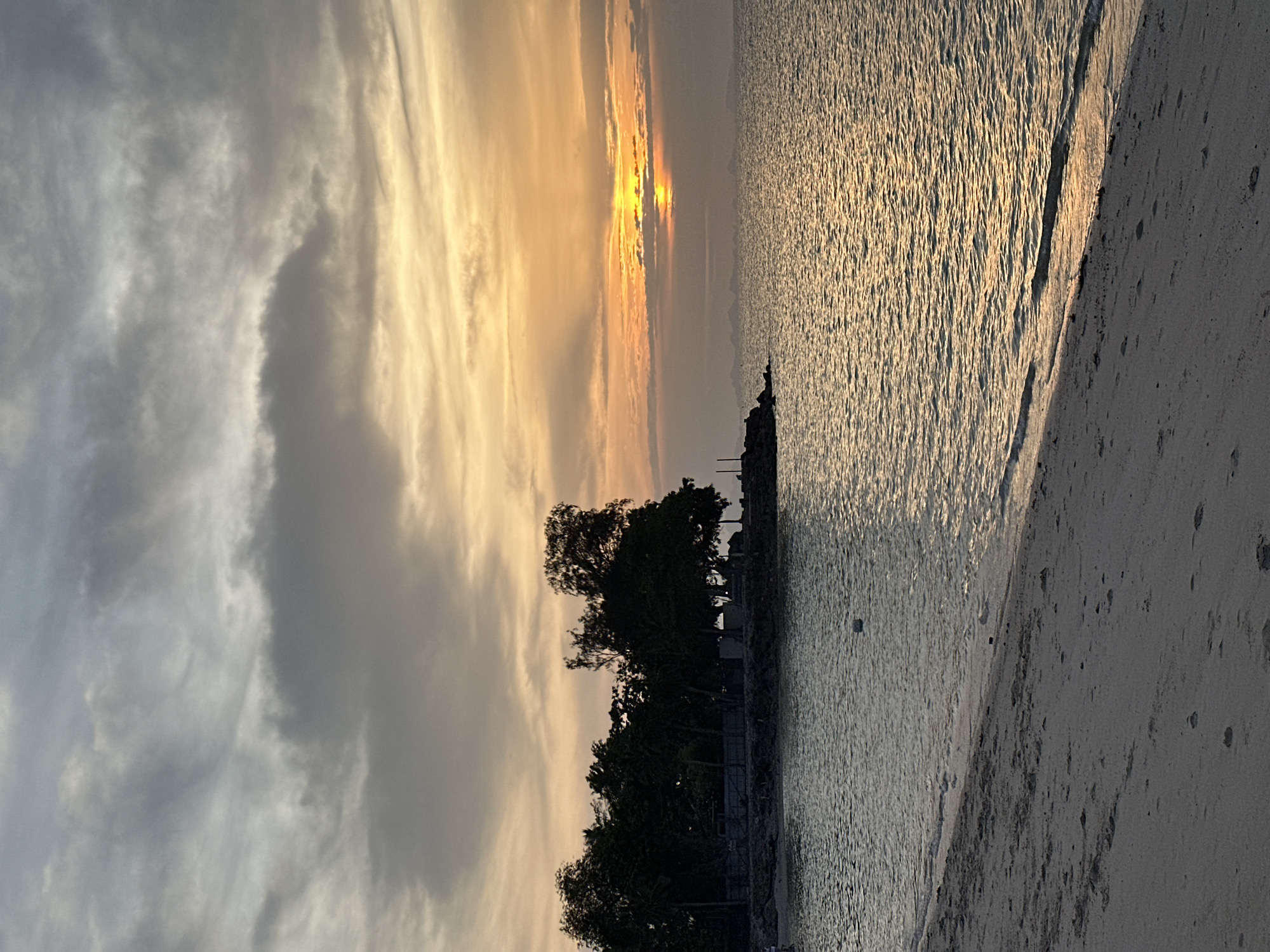 A sunset is visible over the ocean, with a sandy beach in the foreground. The sky is filled with clouds, with streaks of orange and yellow light emanating from the setting sun. The water is calm, with small waves lapping at the shore. There is a line of trees on a rocky outcrop to the left of the image, and a faint outline of islands in the distance. There are no people or animals visible in the image. The scene is peaceful and serene. The location is likely a beach in the Ko Samui District of Thailand.