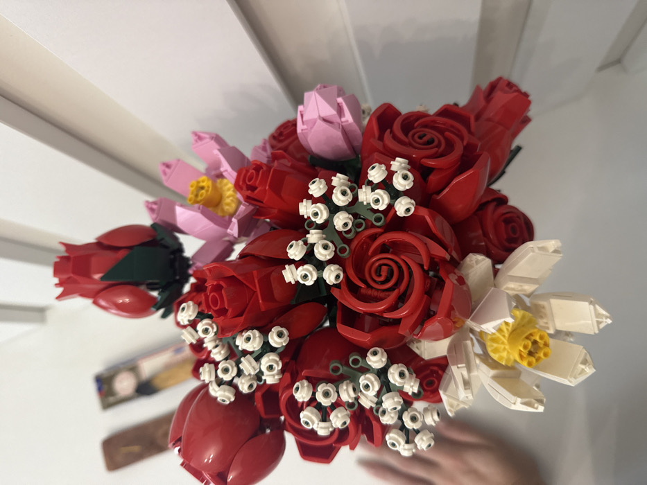 The image displays a close-up of a bouquet composed of artificial flowers constructed from Lego bricks, located in a room in the city of Batumi, Georgia. The bouquet primarily features numerous red roses, some fully open with spiral petals, others in various stages of bloom, and several red buds with green sepals. White 'baby's breath' style clusters, formed from small white studs on green stems, are interspersed throughout. A distinct white daffodil with a prominent yellow trumpet-shaped center is positioned at the bottom left of the arrangement. Towards the upper right, a light pink flower with a yellow center is visible, alongside another pink, more closed bloom. A light-skinned human hand is partially visible and out of focus on the far left, positioned near the bouquet. The background consists of a plain white wall or surface. On the far left, behind the hand and out of focus, two rectangular objects are present: a dark brown bar and a vertically oriented item displaying what appears to be a graphic design or illustration.