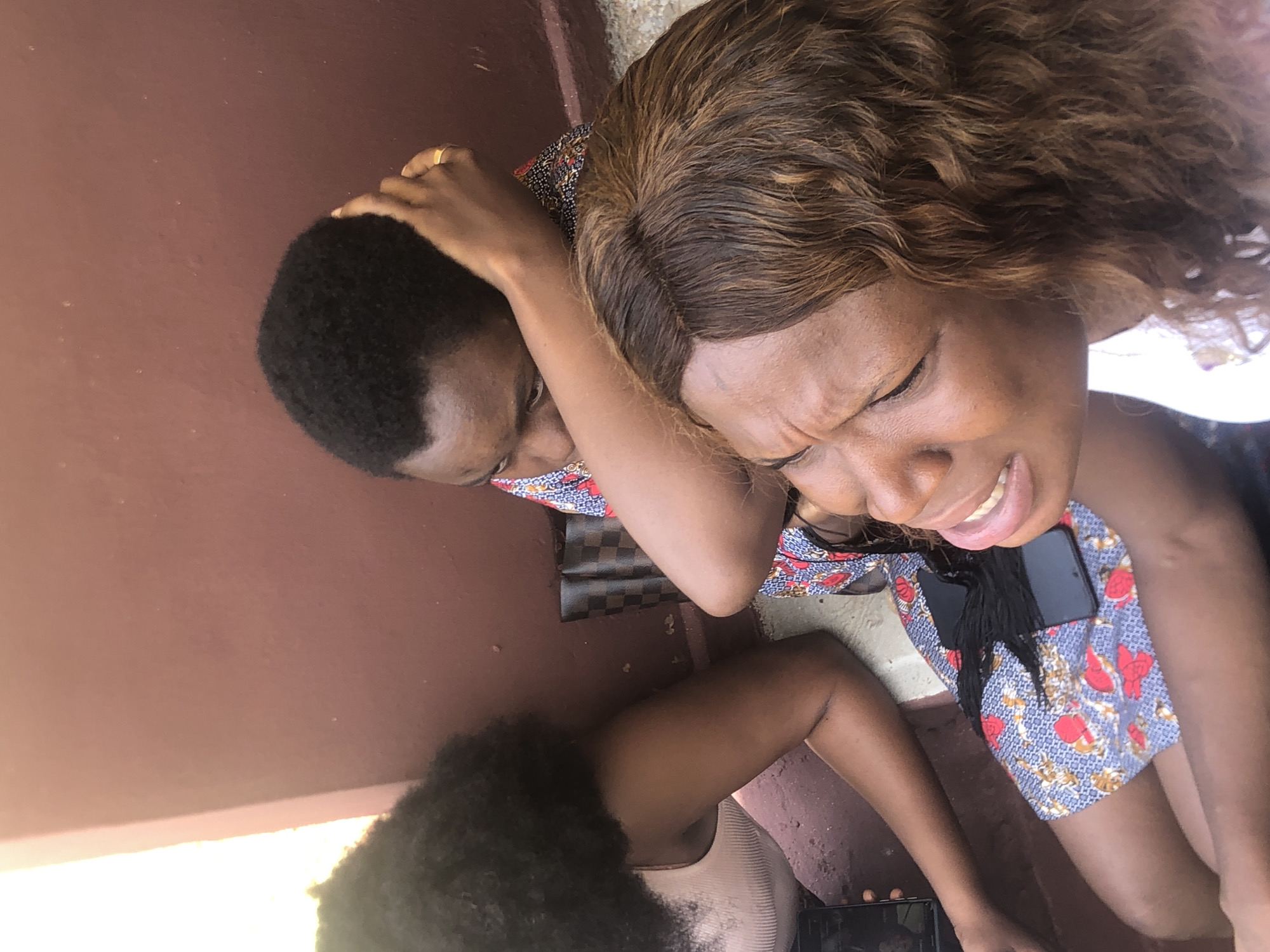 Two Black women are shown close together outdoors, potentially in Ukwu-Abwa, Nigeria. One woman with short, dark hair is holding her head. The other woman, with highlighted brown curly hair, has her eyes closed and appears to be grimacing. Both women are wearing patterned clothing. A third person with curly hair is partly visible in the lower left corner. Mobile phones are visible next to the woman with brown hair. The setting appears to be a paved area next to a reddish-brown wall.