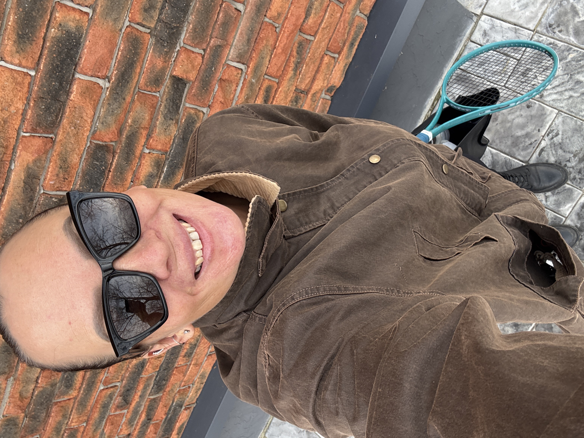 An overhead high-angle selfie depicts an adult female subject standing outdoors in Chaikovsky, Russia. The subject, visible from the chest up, has dark hair pulled back and wears dark, cat-eye style sunglasses that reflect bare tree branches against a bright sky. She presents a wide smile, exposing her teeth. Her left ear displays a silver stud earring on the lobe, a small hoop in the helix, and a decorative cuff on the upper helix. She is dressed in a brown, textured jacket with visible stitching, brass-colored buttons, and a beige corduroy collar. A turquoise-framed tennis racket with white strings rests near her right leg. The background features a brick wall composed of reddish-brown and dark bricks with light mortar. A dark gray ledge is positioned below the brick wall. The ground beneath the subject consists of light gray square tiles.