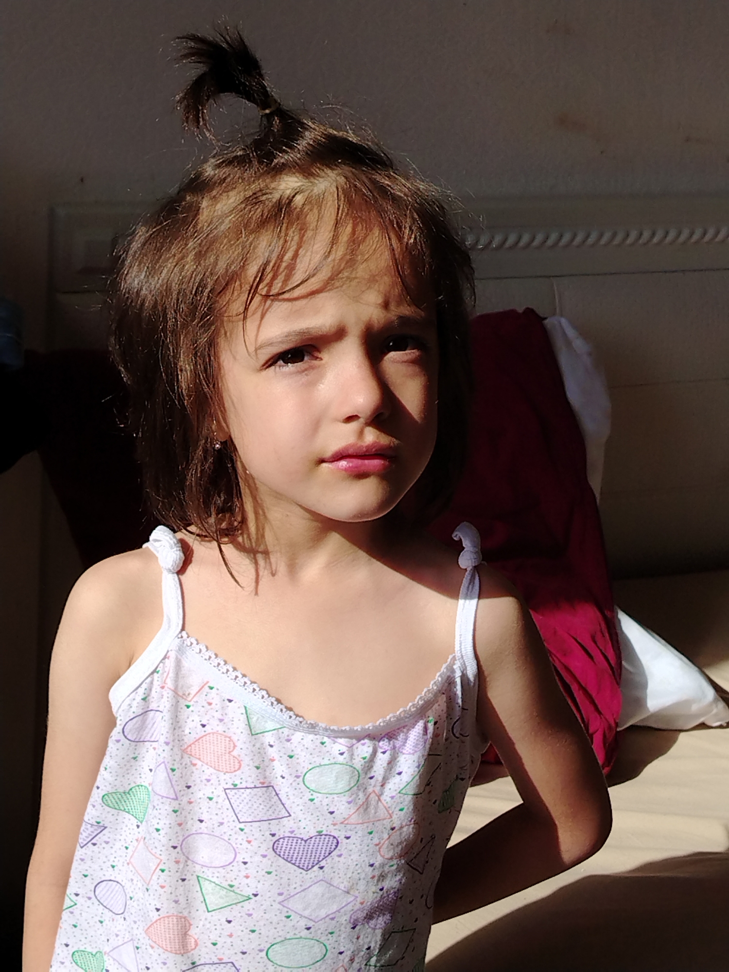 A young Caucasian girl with brown hair pulled into a messy ponytail is sitting against a headboard, likely in a bedroom. She is wearing a white tank top with a colorful geometric pattern of hearts, triangles, and circles. The lighting suggests it is daytime, with strong sunlight casting shadows on her face and body, as well as on the wall behind her. The background shows a bed with a red pillow. The location is not specified in the image.