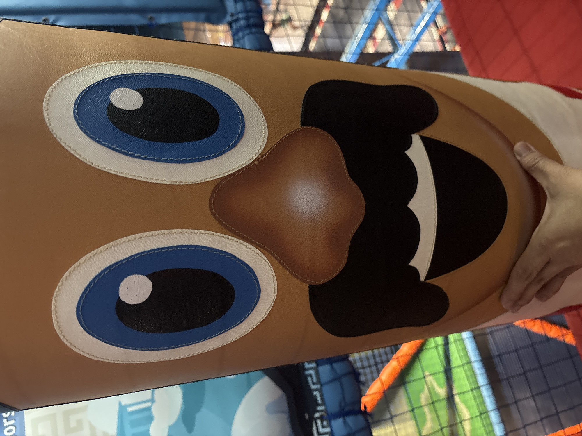 The image shows a close-up view of a large, cylindrical, cartoon-like character's face, likely part of an inflatable structure or play equipment. The face is predominantly tan, with large blue eyes outlined in white and black pupils. A brown, cartoonish nose is positioned between the eyes, and below it is a large black mouth with a white smile. The character appears to have a black mustache. A human hand is visible at the bottom of the frame, touching the character's lower face. The background suggests an indoor play area, with glimpses of blue netting, orange bars, and a red surface, possibly a soft play mat. The location is not specified within the image itself.