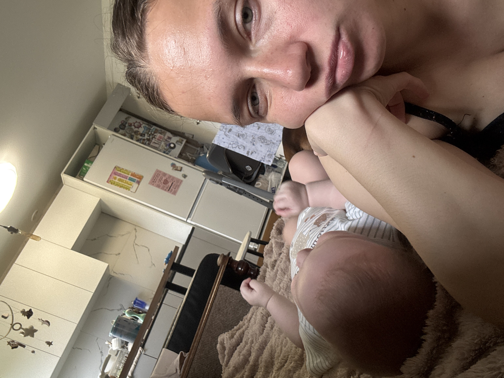 A selfie-style photograph shows a woman indoors in a kitchen-like environment. The woman is fair-skinned, with light-colored eyes and her hair pulled back. She rests her face on her hand, seemingly looking at the camera. Part of her chest is visible with the strap of a black garment. A baby is lying near her. The baby has a light complexion, and a white and light grey striped onesie is visible. A beige textured blanket is underneath them both. In the background, there are white cabinets and appliances, including a refrigerator. There is a mobile with hanging elements near the cabinets. The location is implied to be in Раздолье, Russia.