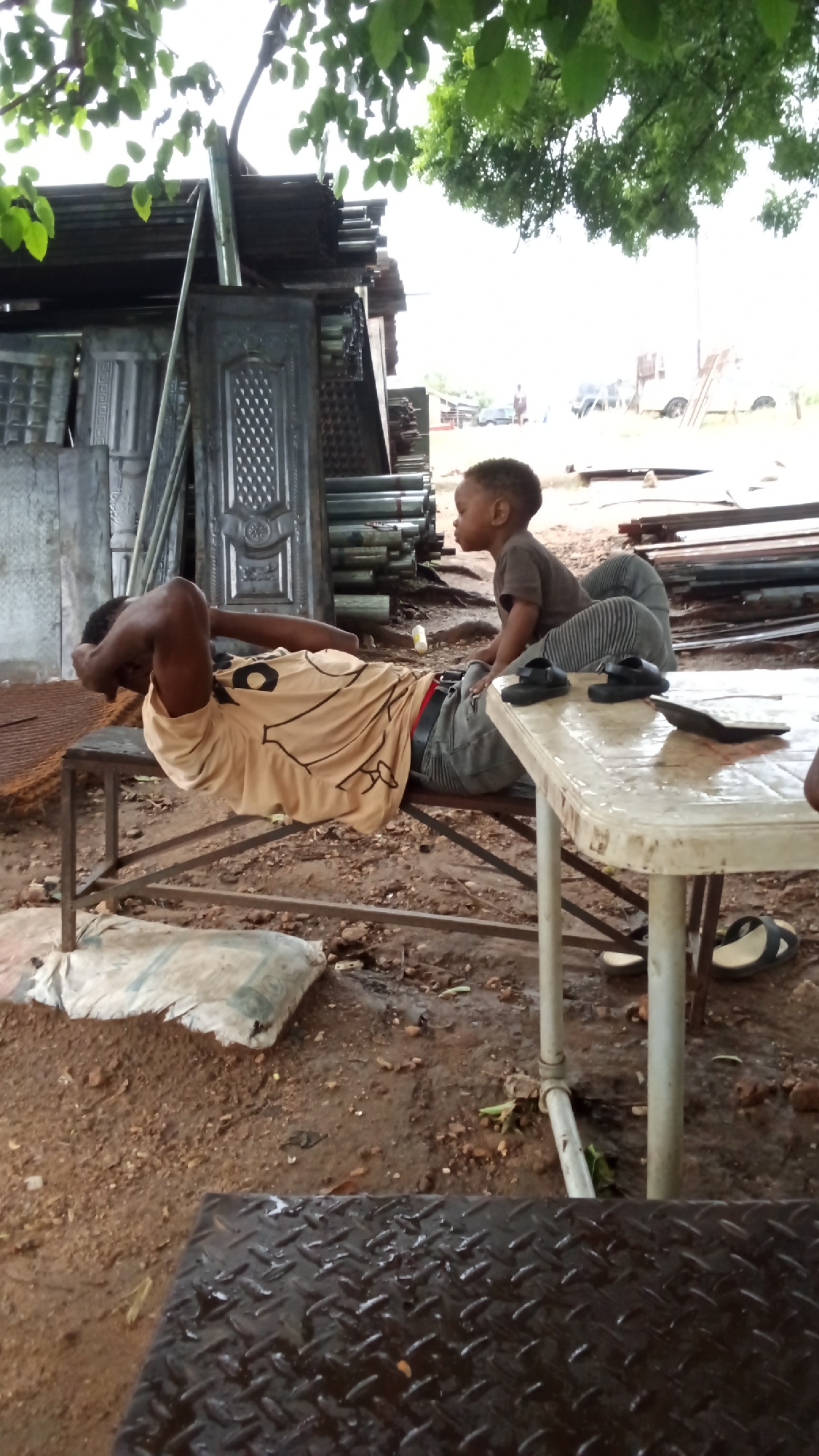In Zango-ndaji, Nigeria, a man with dark skin, wearing a light brown t-shirt and dark pants, lies on a metal bench, his head supported by his hands. He appears to be performing sit-ups. Beside him, a young child with dark skin, wearing a gray t-shirt and striped pants, sits on a white plastic table, looking to the left. The environment is outdoors, with a backdrop of stacked metal doors and pipes. The ground is dirt and appears wet. A white bag is on the ground near the bench.