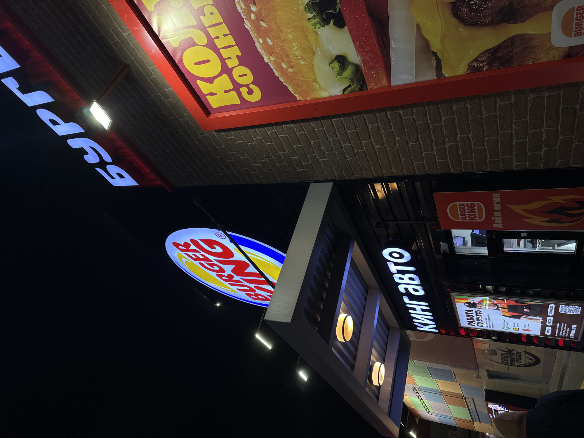 The image depicts a Burger King restaurant in Rostov-on-Don, Russia at night. The photograph is taken at an angle, with the Burger King logo and "кинг авто" (King Auto) sign prominently displayed. Other signage includes advertisements for burgers and job openings. The restaurant's exterior has a modern design with decorative lighting and brickwork. The environment is urban, suggesting a bustling location with other commercial buildings visible in the background. A person's shoulder is visible in the lower right corner.