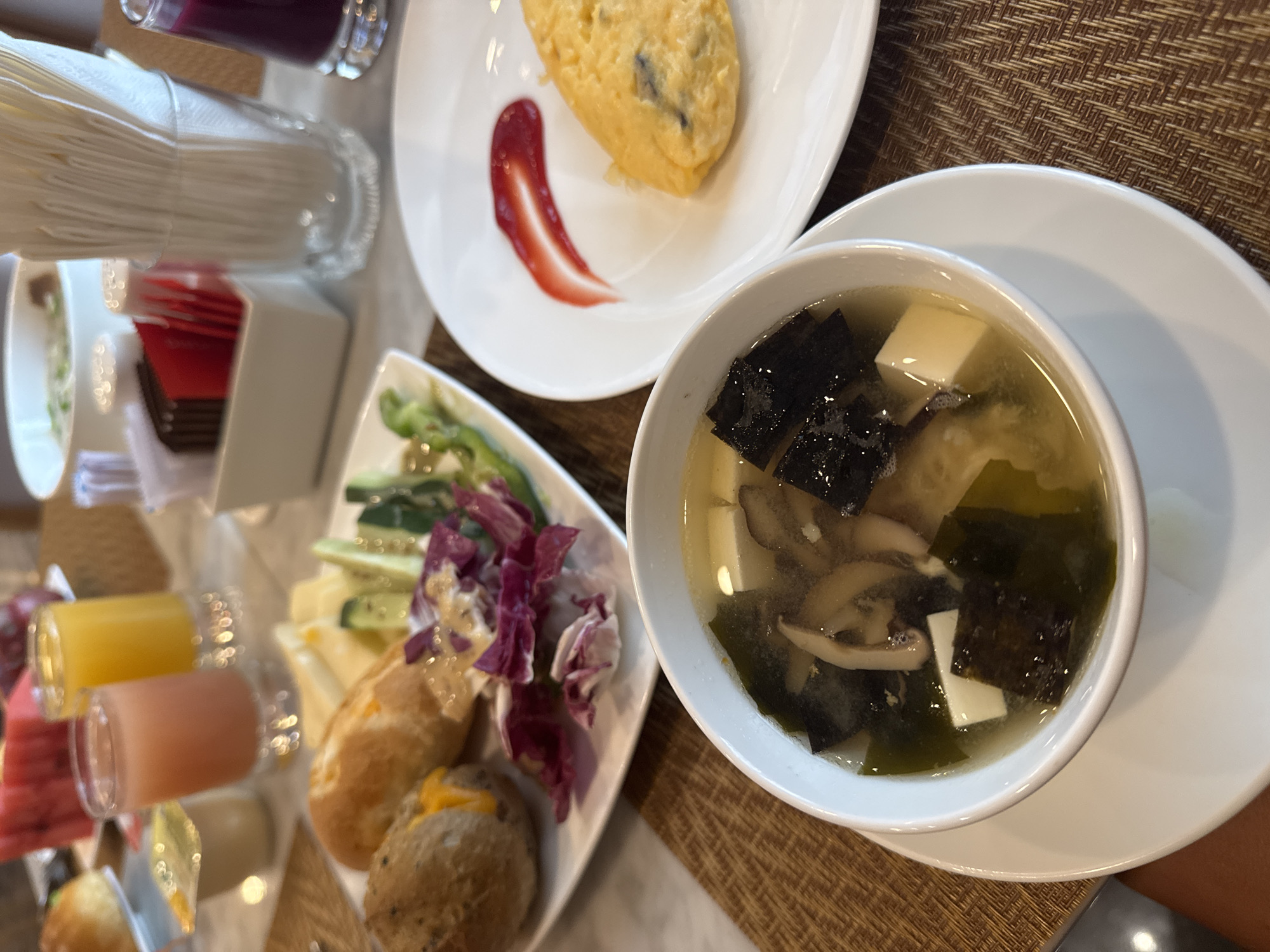 A top-down shot reveals a breakfast spread, likely in Đà Nẵng, Vietnam given the presence of Miso Soup, on a woven table surface. The meal consists of an omelet with ketchup on a white plate, a plate containing assorted vegetables, cheeses, bread rolls, and small glasses of juice and sliced watermelon. The bowl of Miso Soup contains seaweed, tofu, and mushrooms. Utensils are set to the side. A dispenser holding napkins sits to the left. The lighting suggests daytime.