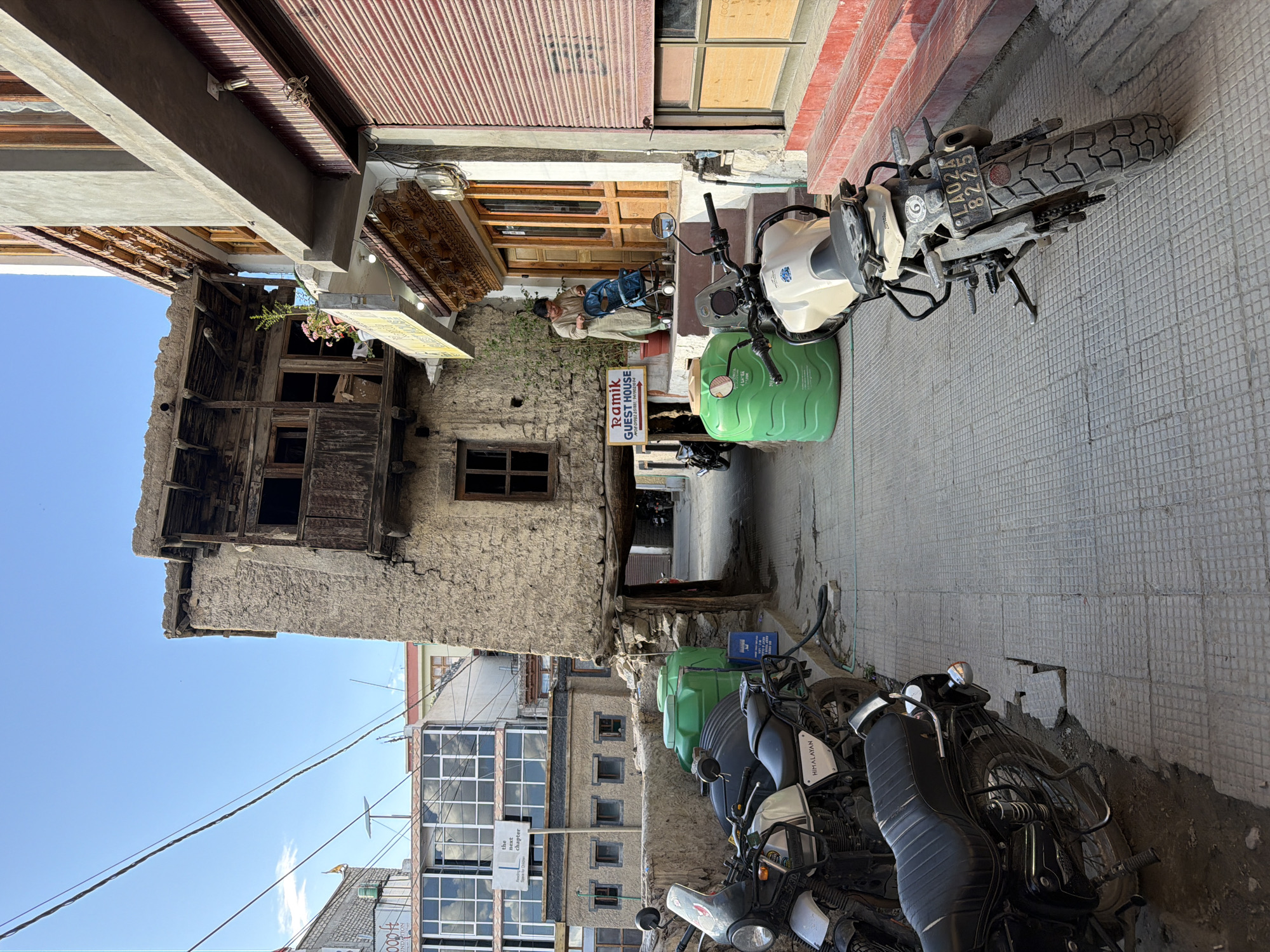 This is a street-level, eye-level photograph taken in Leh, India, showcasing a narrow alleyway lined with traditional Ladakhi architecture. The scene is dominated by a rustic, multi-story building constructed with mud and stone, featuring weathered wooden balconies and window frames, suggesting significant age. To the right, a more modern building with a painted facade and traditional wooden doors and window detailing is visible. A woman in traditional clothing is pushing a blue stroller up a set of red-painted steps leading to the entrance of the building on the right. Parked along the left side of the alley are several motorcycles. Two large green water tanks are positioned near the motorcycles. In the background, a modern building with large glass windows and a sign that reads "the next chapter" is present, contrasting with the older structures. The ground is paved with square tiles. The sky is a clear blue with a few wispy clouds. The overall impression is of a bustling yet historically rich street in Leh.