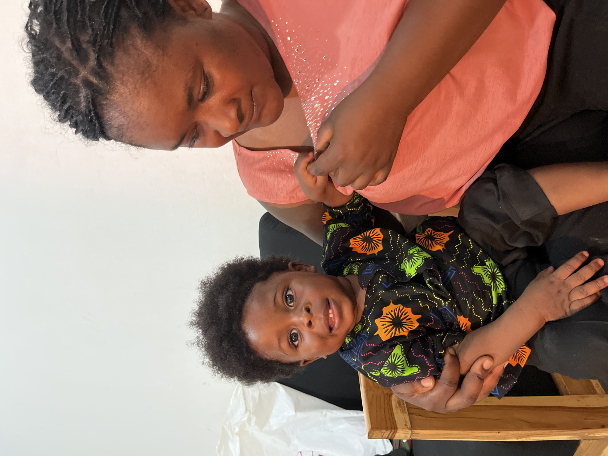 In Gura Top, Nigeria, a woman with braided hair is seen in a close-up shot, holding a young child. The woman is wearing a pink, sparkly top and black pants, and she is looking down at the child with a gentle expression. The child, who has a mop of dark, curly hair, is wearing a black shirt with a colorful, floral pattern. The child is looking directly at the camera with wide, curious eyes and a slight smile.
The image appears to be taken indoors, with a plain white wall as the background. The lighting suggests it is daytime. The interaction between the woman and the child conveys a sense of warmth and affection. The focus is on the faces and hands of the two subjects, highlighting their connection. No visible text is present in the image.