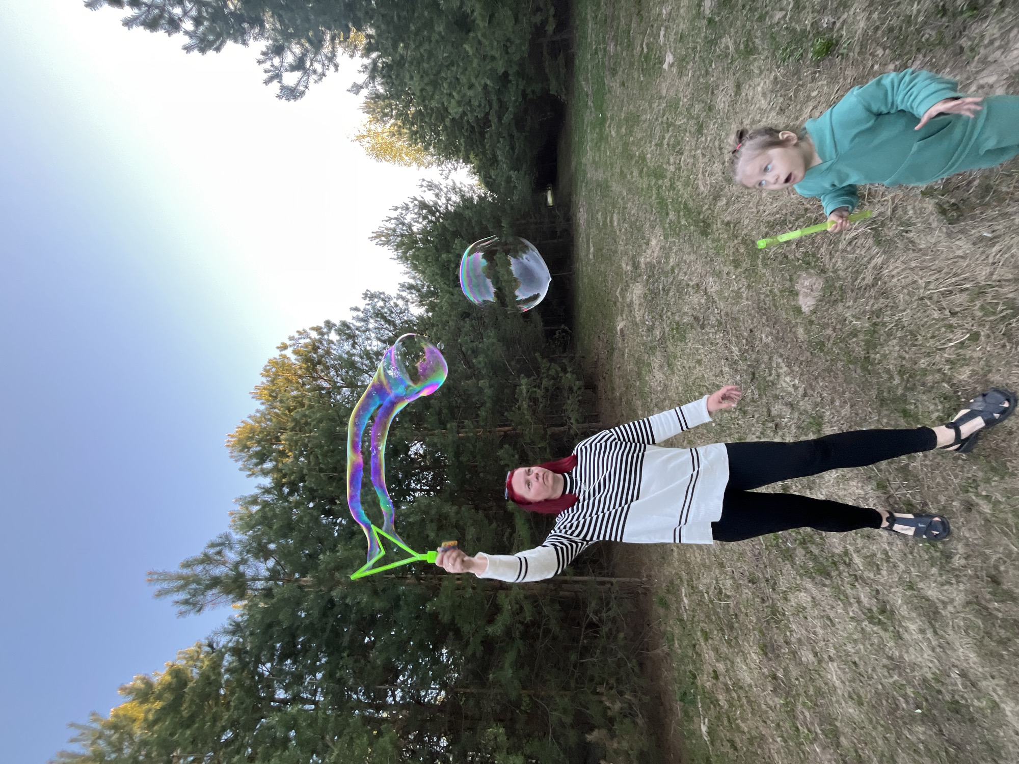 A woman with red hair, wearing a white and black striped shirt and black leggings, is holding a green bubble wand and creating a large, rainbow-colored bubble. Another child, wearing a green outfit, is running behind her, also holding a green bubble wand. There are two bubbles in the air. The scene takes place in a grassy area surrounded by trees, possibly a park. The sky is visible in the background. This appears to be a recreational area possibly in the city of Chaikovsky, Russia.