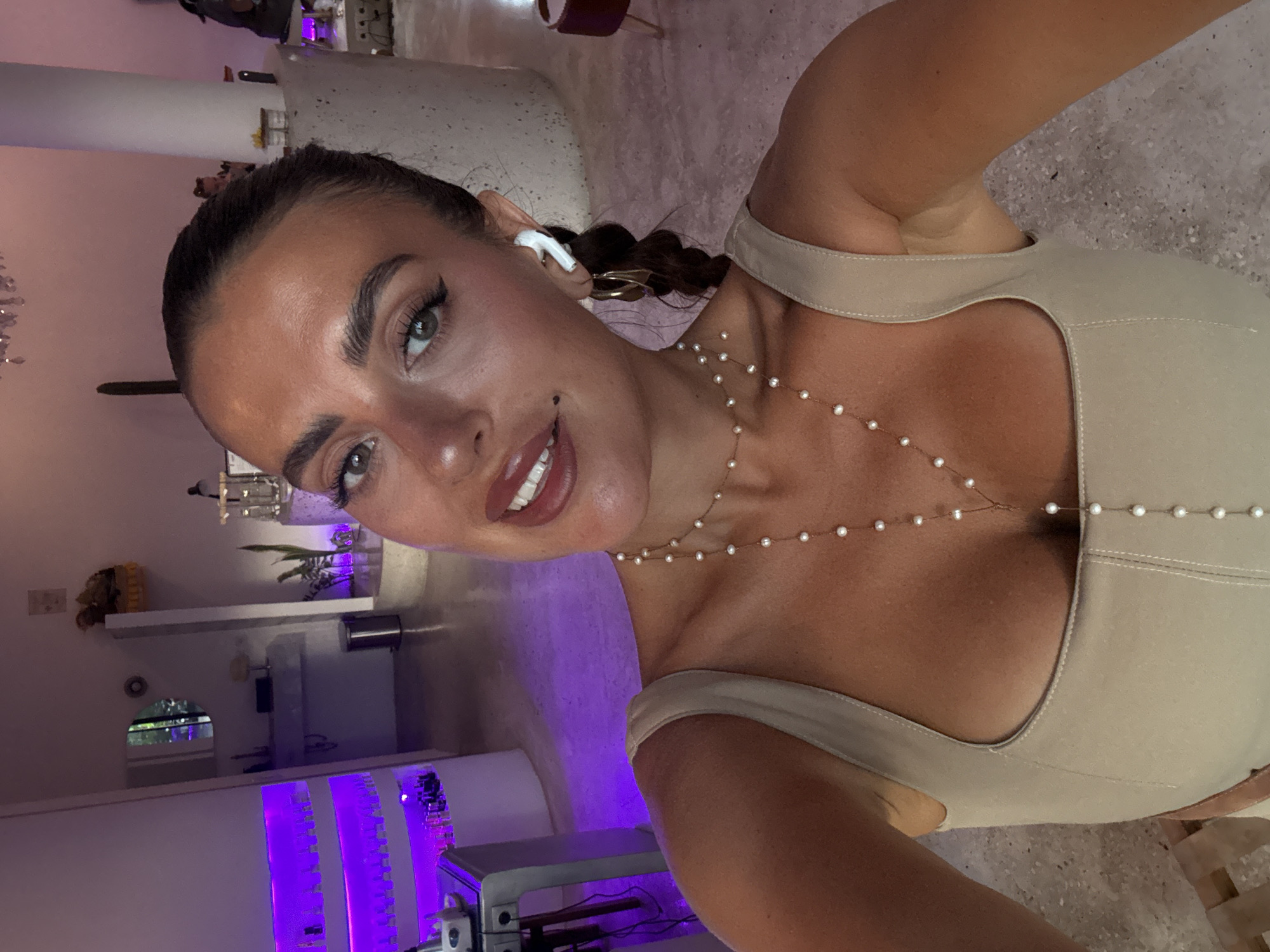A fair-skinned female poses in a tan top, wearing an earbud and a pearl body chain. She has brown hair and makeup, including dark eyeliner and lipstick. The location is identifiable as North Kuta, Indonesia based on the architectural style, and the purple lighting in the background suggests a modern interior design.