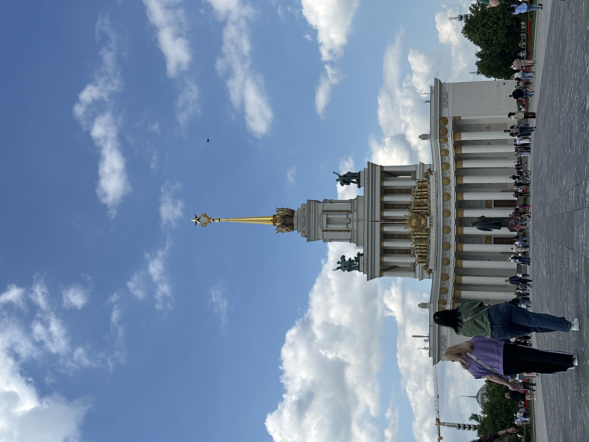 The image captures a view of the VDNKh Main Pavilion in Moscow, Russia, on a partly cloudy day. The architectural structure features a tall spire topped with a Soviet star, flanked by elaborate facades and statues. Numerous people are present on the paved plaza in front of the pavilion, some walking or standing. The foreground includes two figures, one in a purple shirt and the other in a green jacket. The sky is blue with scattered white clouds.