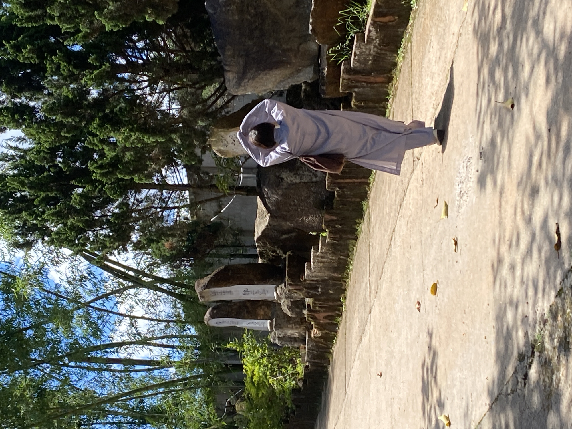 A person dressed in a light purple robe is standing with their back to the camera, their hands clasped above their head. They are positioned on a concrete path in what appears to be a park or garden setting. The path is bordered by a rustic stone wall and lush green foliage. In the background, large rocks and trees are visible, with sunlight casting shadows on the ground. The scene is outdoors, with a bright sky peeking through the tree branches. A small brown pouch is hanging from the person's waist. There are some pieces of writing on the rocks in the background that are not legible in the image. The overall impression is one of peace and contemplation.